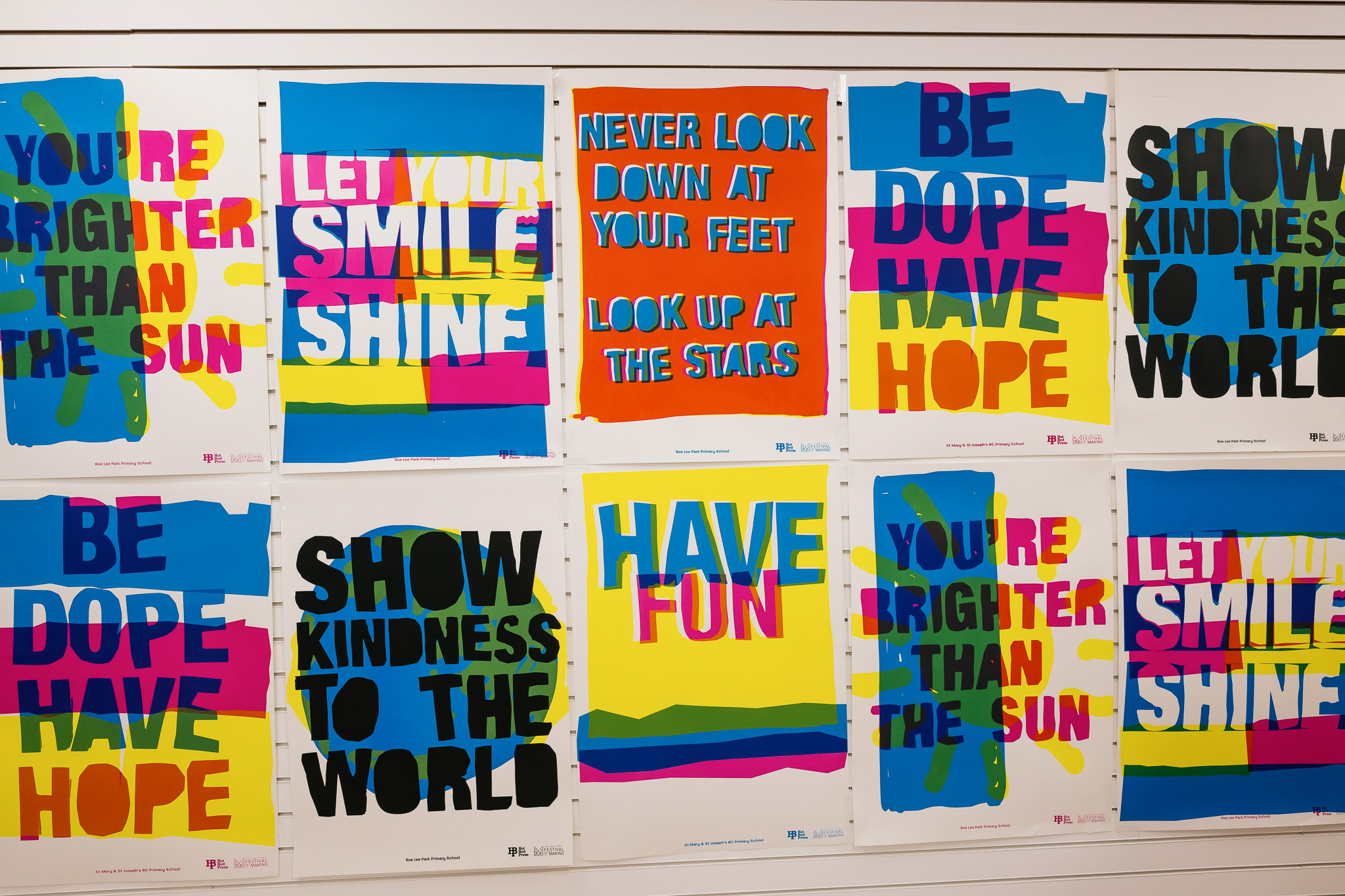 Nine colourful posters with bold, overlapping text featuring positive messages like “LET YOUR SMILE SHINE,” “NEVER LOOK DOWN AT YOUR FEET LOOK UP AT THE STARS,” and “SHOW KINDNESS TO THE WORLD” displayed on a white wall.