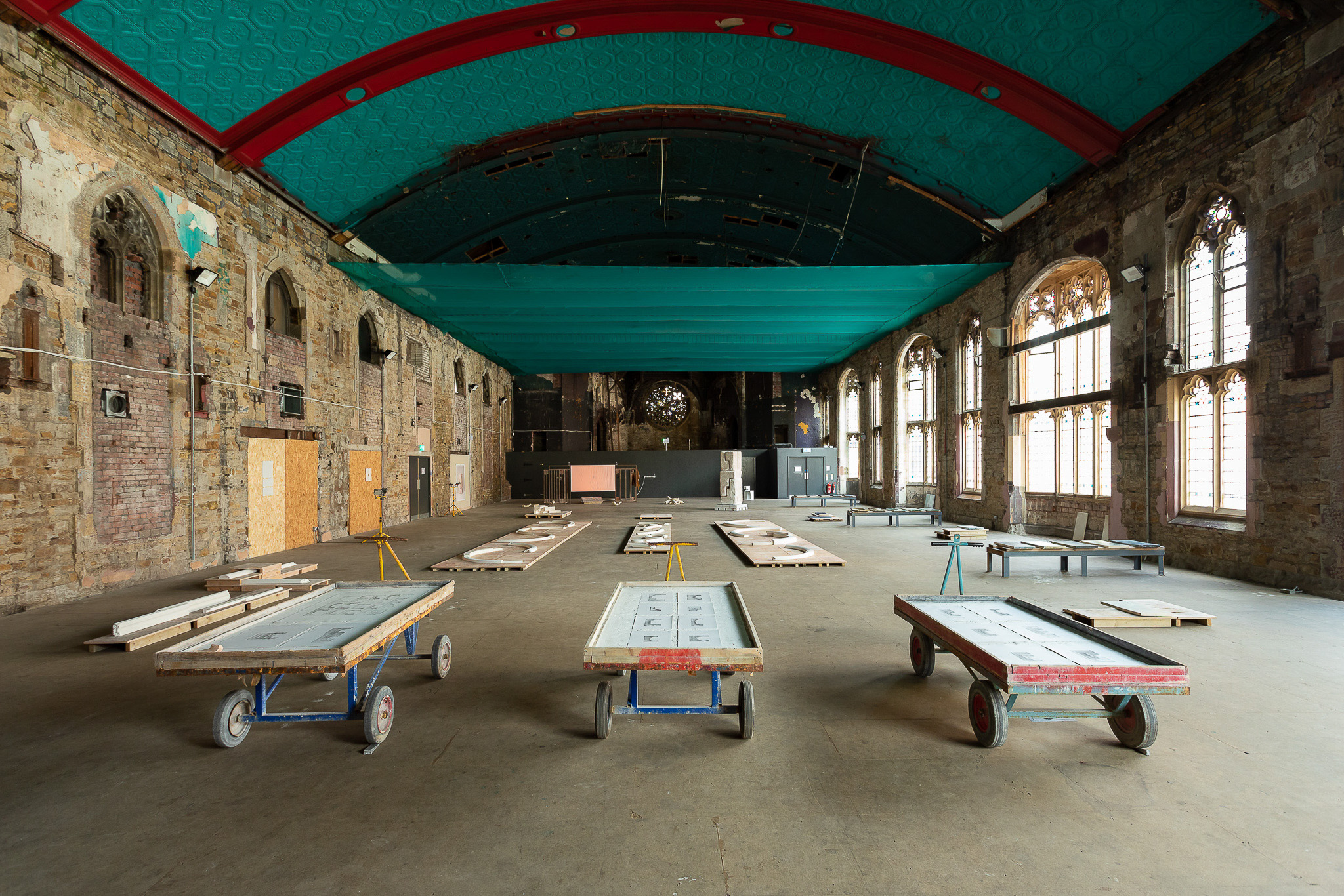 A large, heritage hall with a turquoise ceiling and exposed brick walls. Three wooden trolleys are in the foreground with artwork on top, and pallets and sculptural artworks are placed around the spacious, partially restored interior.