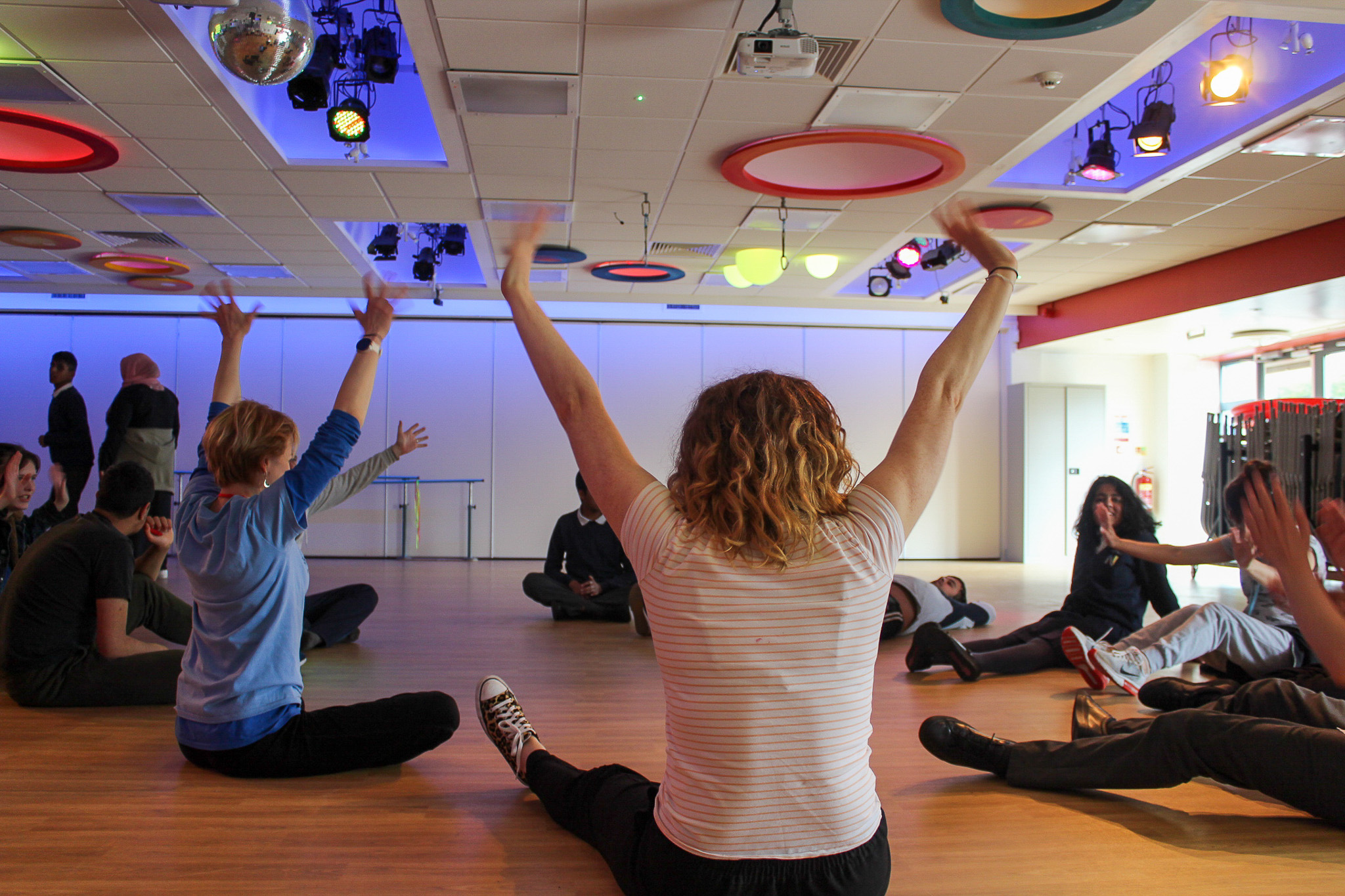 People sit in a circle on a wooden floor, raising arms in a brightly lit room with colourful ceiling lights and a disco ball.