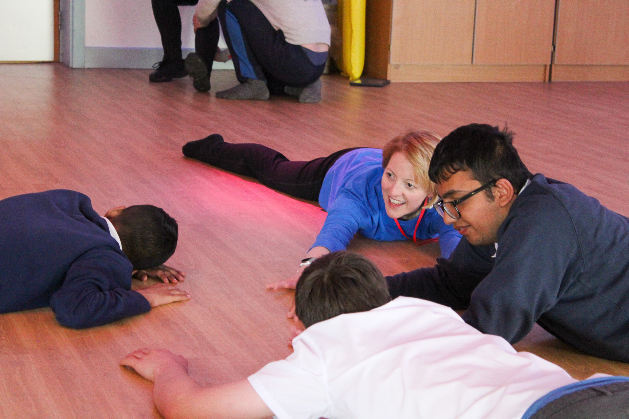 Children and a teacher lie on a wooden floor, smiling and engaging in a playful activity together in a classroom setting.