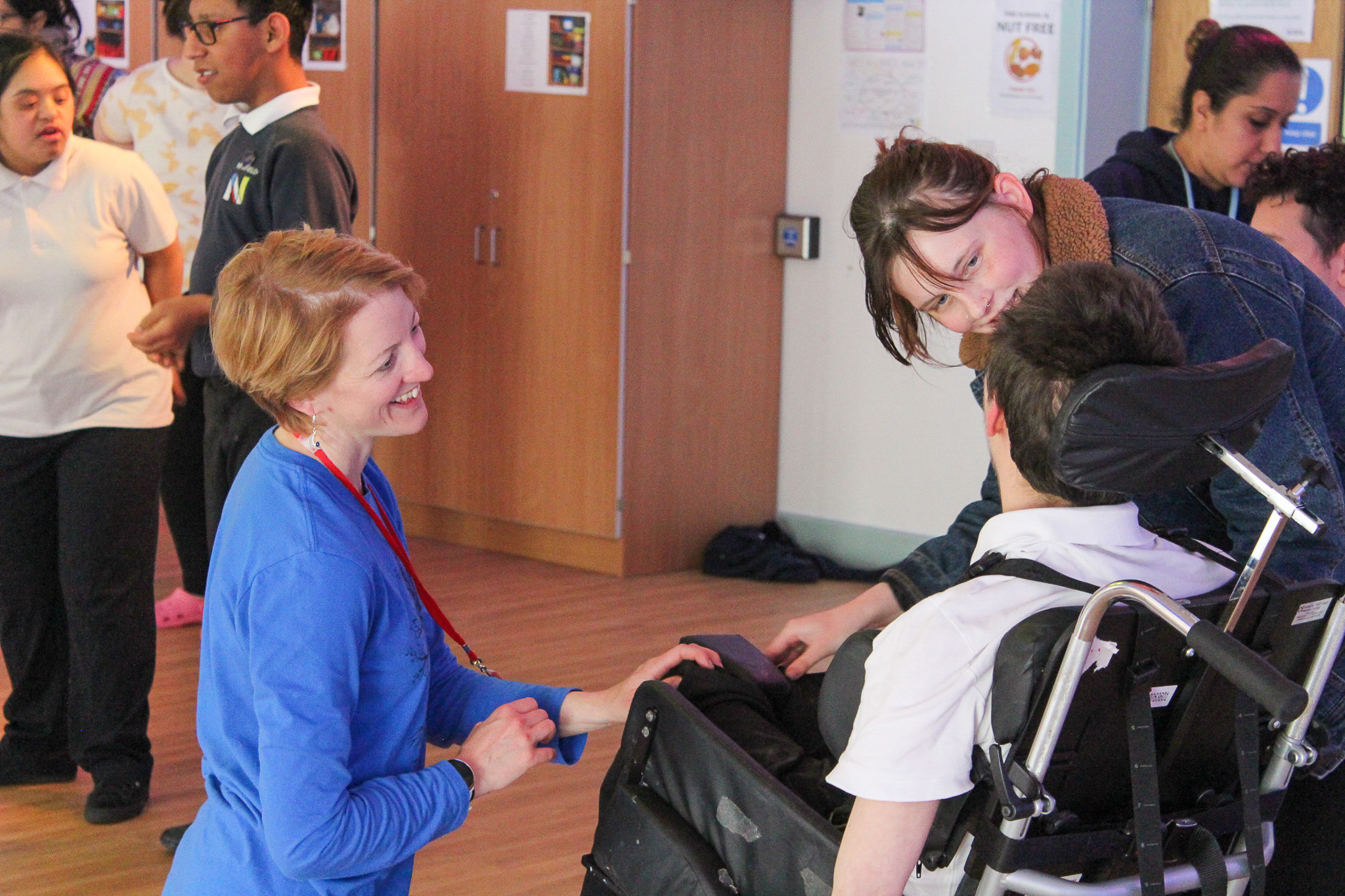 A teacher in blue interacts warmly with a student in a wheelchair, while others engage in the background of a classroom setting.