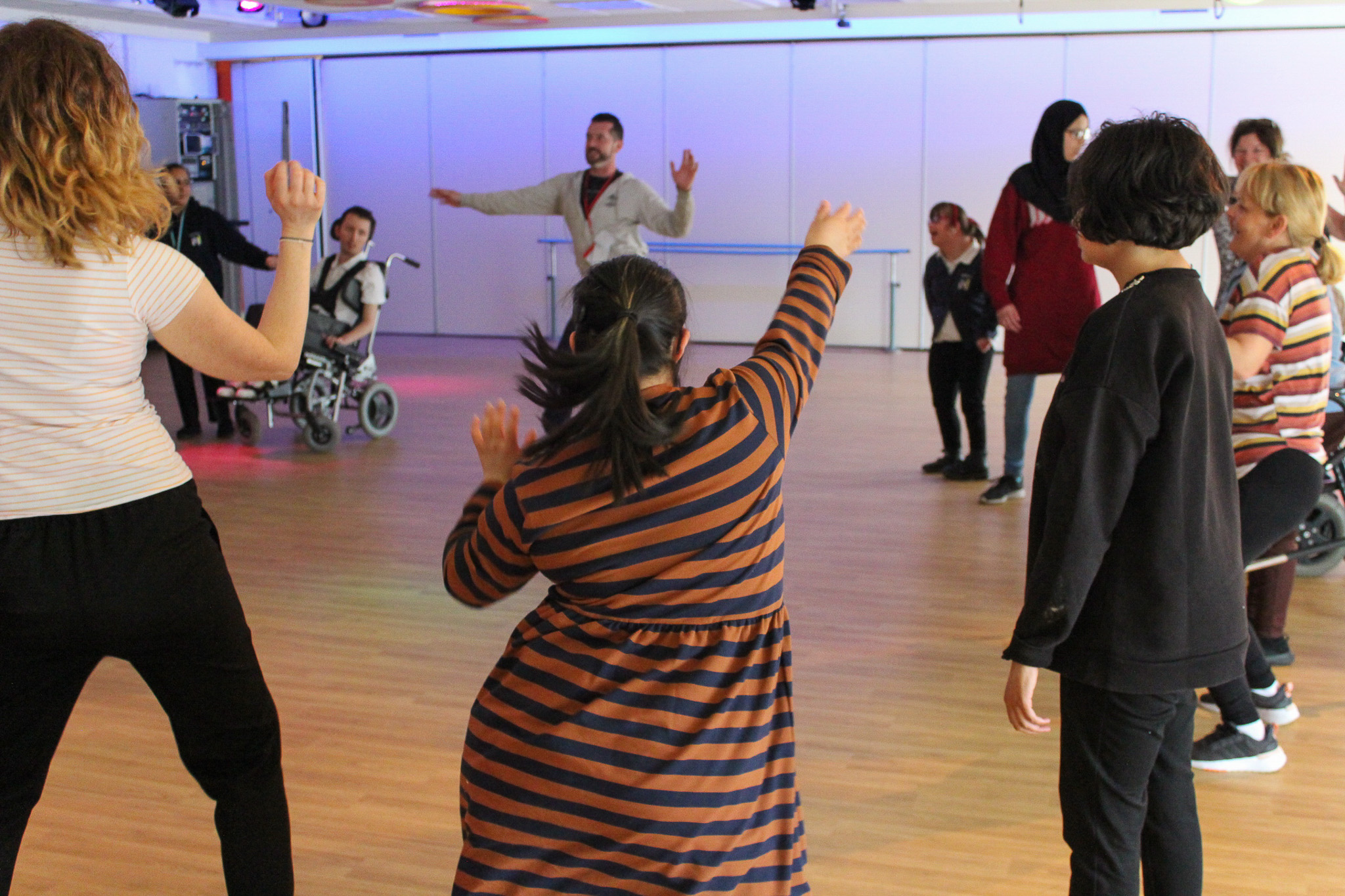 A group of people, including individuals in wheelchairs, dance and enjoy themselves in a spacious, brightly-lit room with wooden floors and white walls. Everyone appears to be engaged and having fun together.