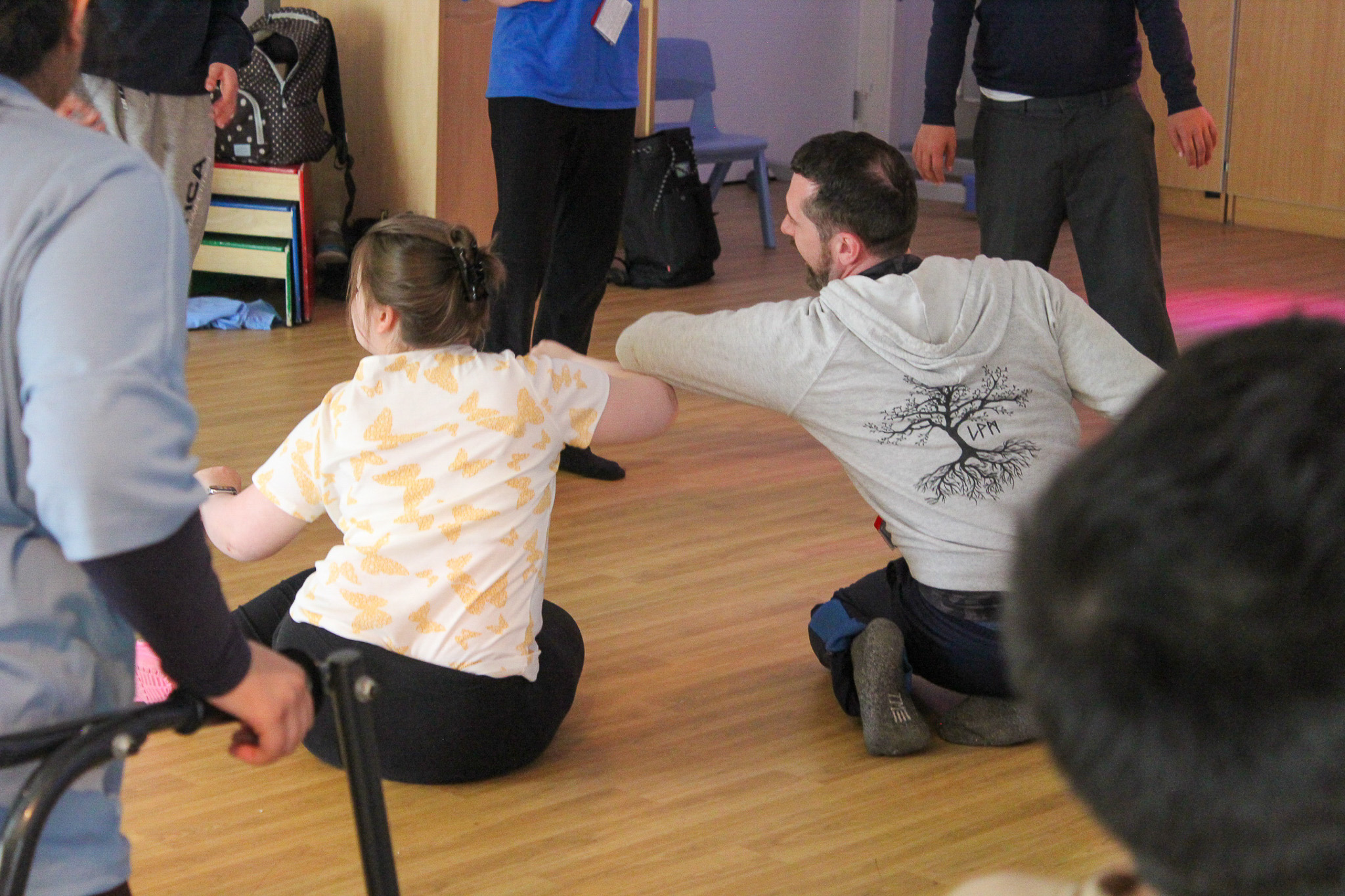 People in a circle, two sitting on the floor, engaged in a group dance activity on a wooden floor.