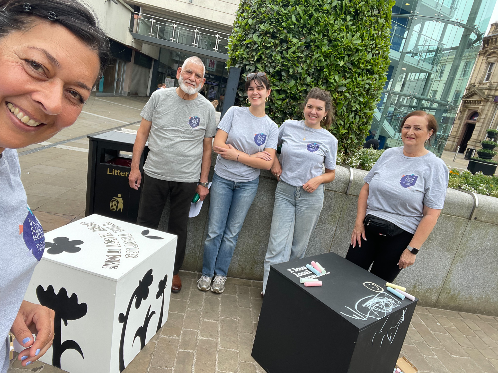 Five people wearing matching grey shirts stand outdoors beside decorated cubes and chalk, smiling at the camera. There are buildings and greenery in the background.