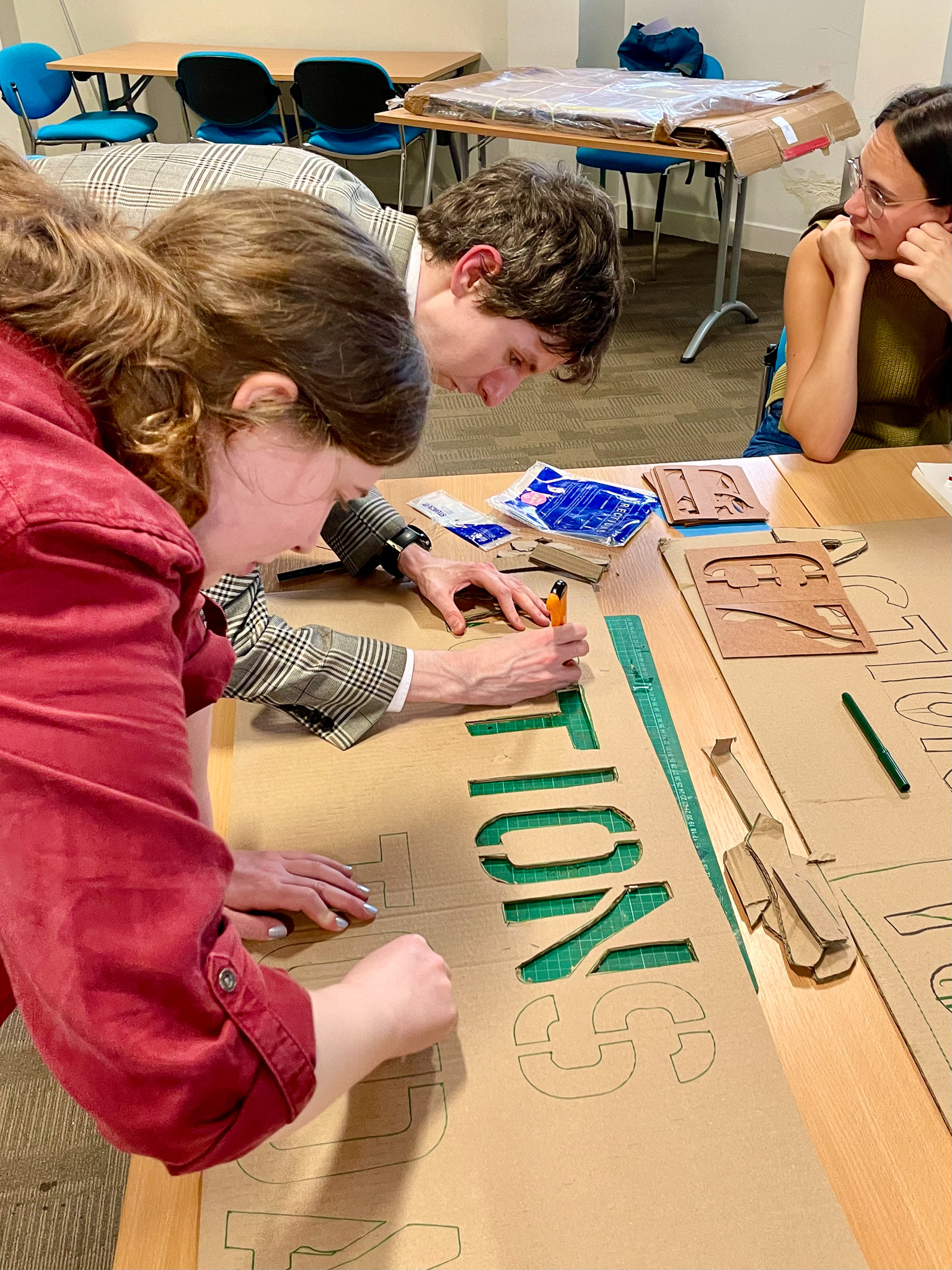 Three people collaborate on a cardboard stencil project at a table, focusing intently on cutting out large letters.