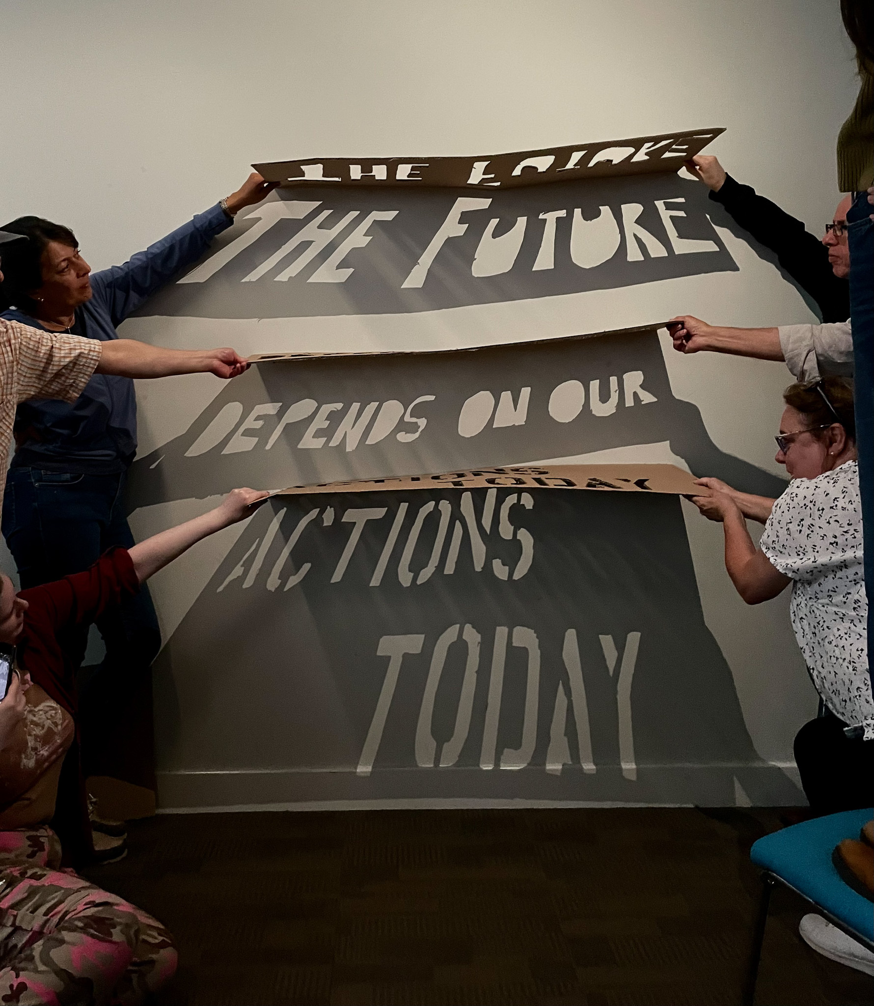 Six people hold large cut-out banners against a wall, displaying the message: THE FUTURE DEPENDS ON OUR ACTIONS TODAY. The message casts shadows on the wall, creating a dramatic visual effect.