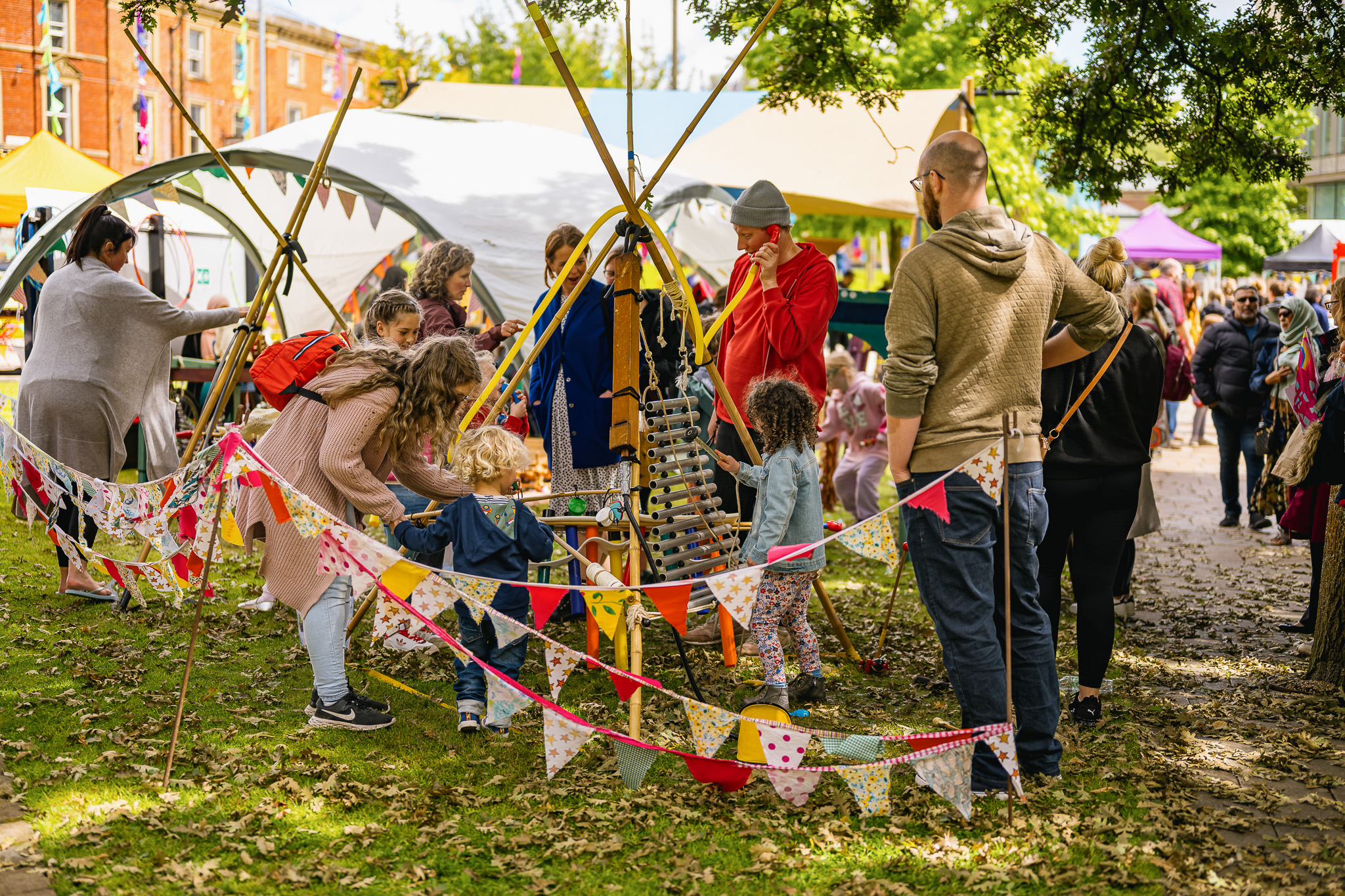 A group of adults and children gather around a small, colourful outdoor play area decorated with bunting at a community fete, with tents, trees, and people visible in the background.