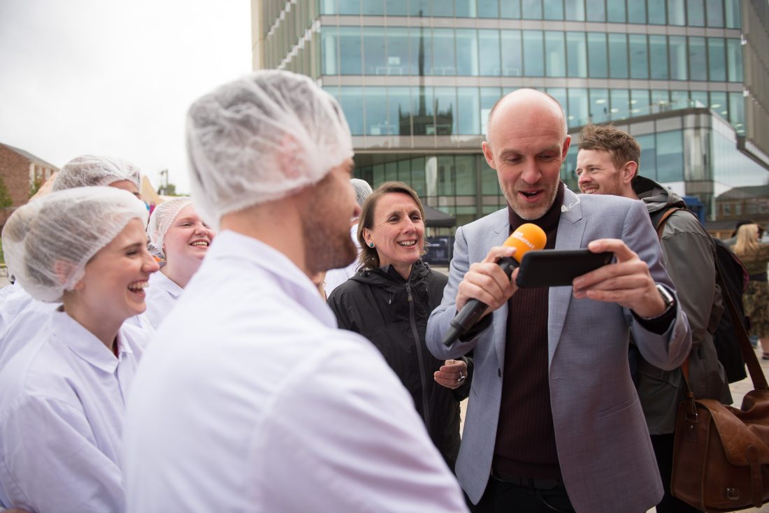 A man in a suit holds a microphone and mobile phone, speaking to a group of smiling people in white coats and hairnets outdoors near a modern glass building.