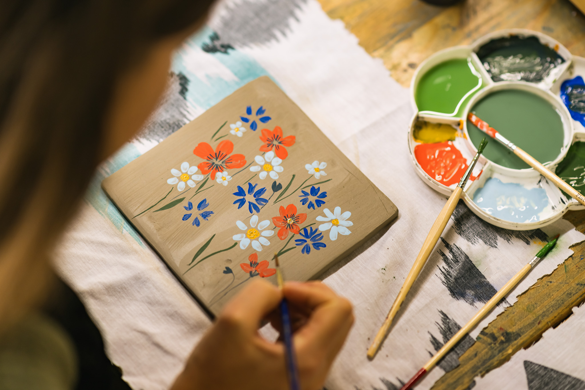 A person paints colourful red, blue, and white flowers on a rectangular piece of wood, with a paint palette and brushes on a table nearby.