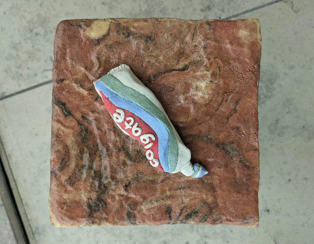 A sculpture of a crumpled Colgate toothpaste tube rests on a textured, square, stone-like surface with reddish and brown marbled patterns. The photo is taken from above.