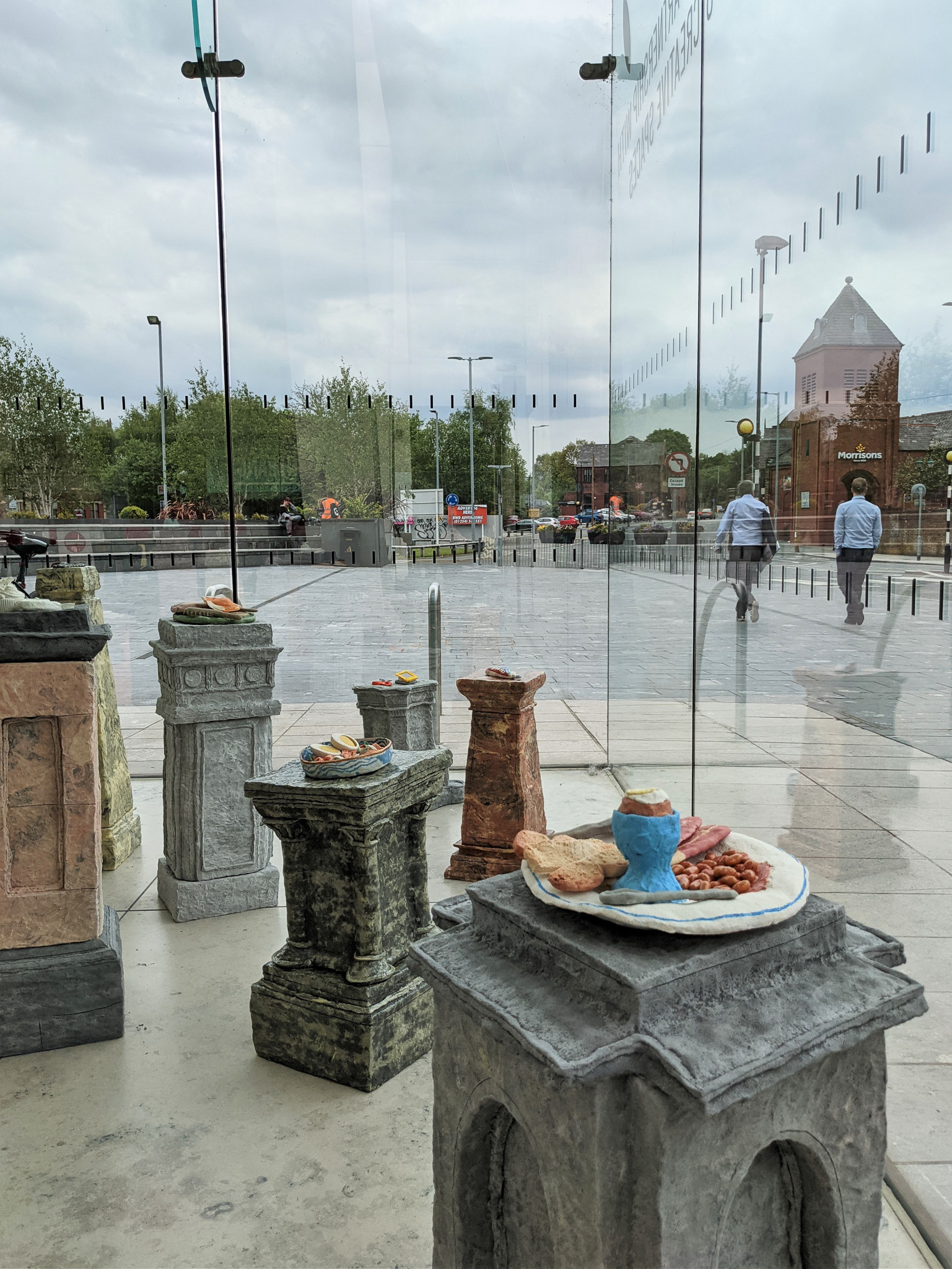 Plates of food sculptures are displayed on stone-like plinths inside a glass case outdoors, with people walking in the background and buildings visible under a cloudy sky.