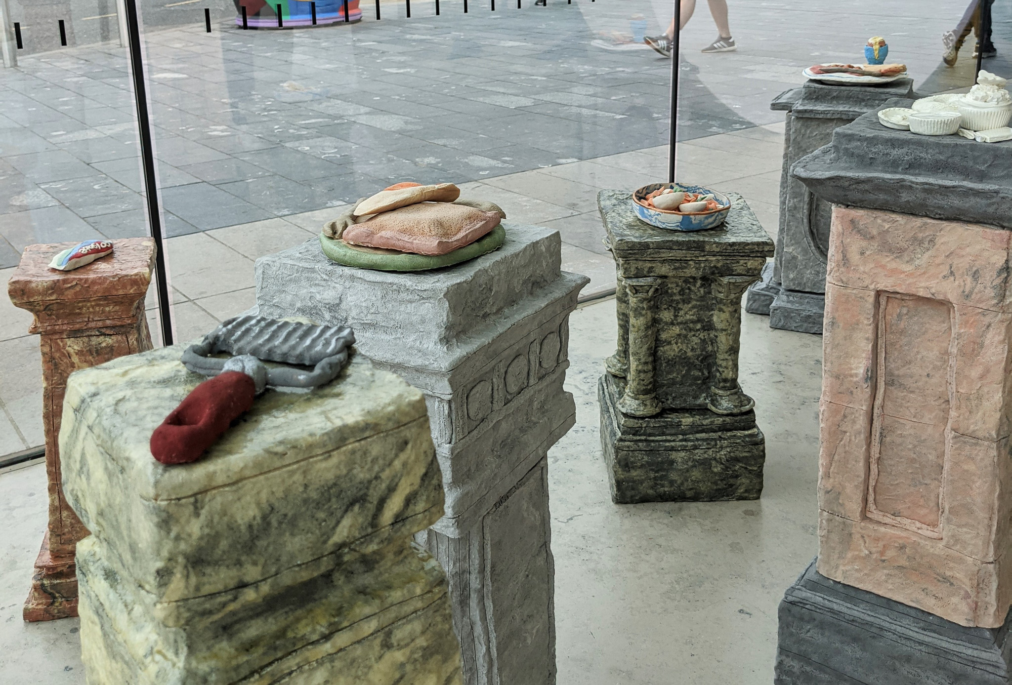 Sculpted stone plinths display colourful, realistic food and utensil sculptures inside a glass enclosure; outside, people walk on a paved pavement.