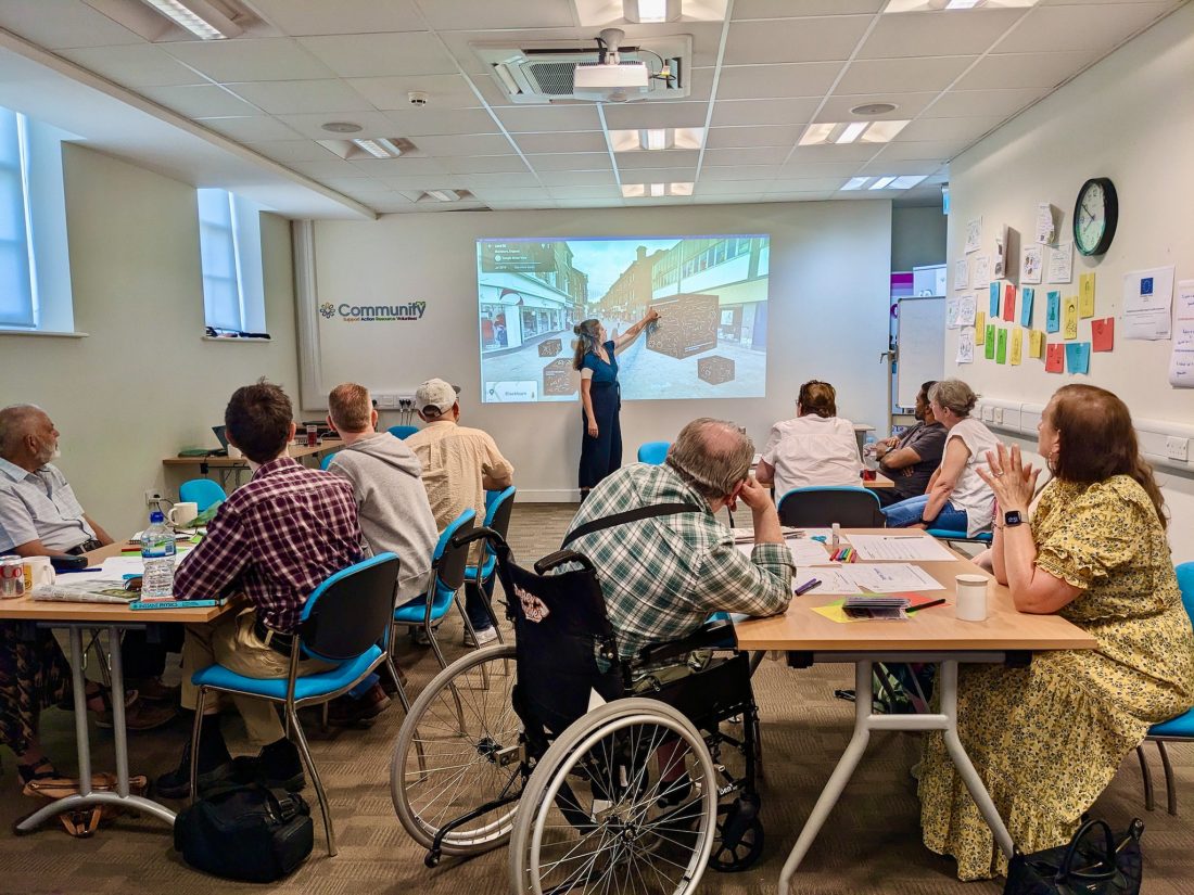 A group of adults, including a person in a wheelchair, sit at tables in a classroom, listening to a woman presenting with a projector screen at the front displaying images of a community space.