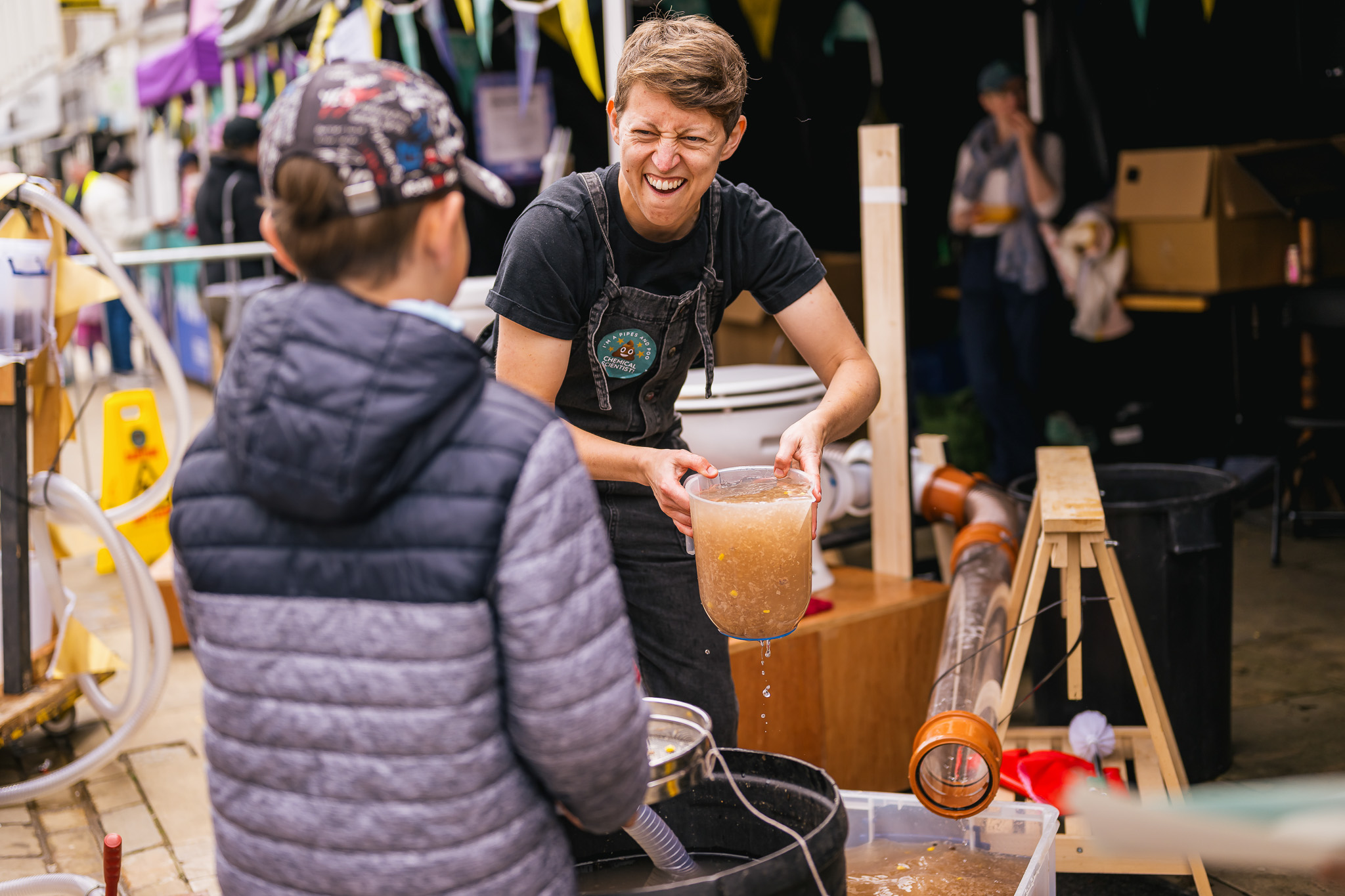 A person smiles while pouring a jug of murky liquid at an outdoor event, interacting with a child in a cap.