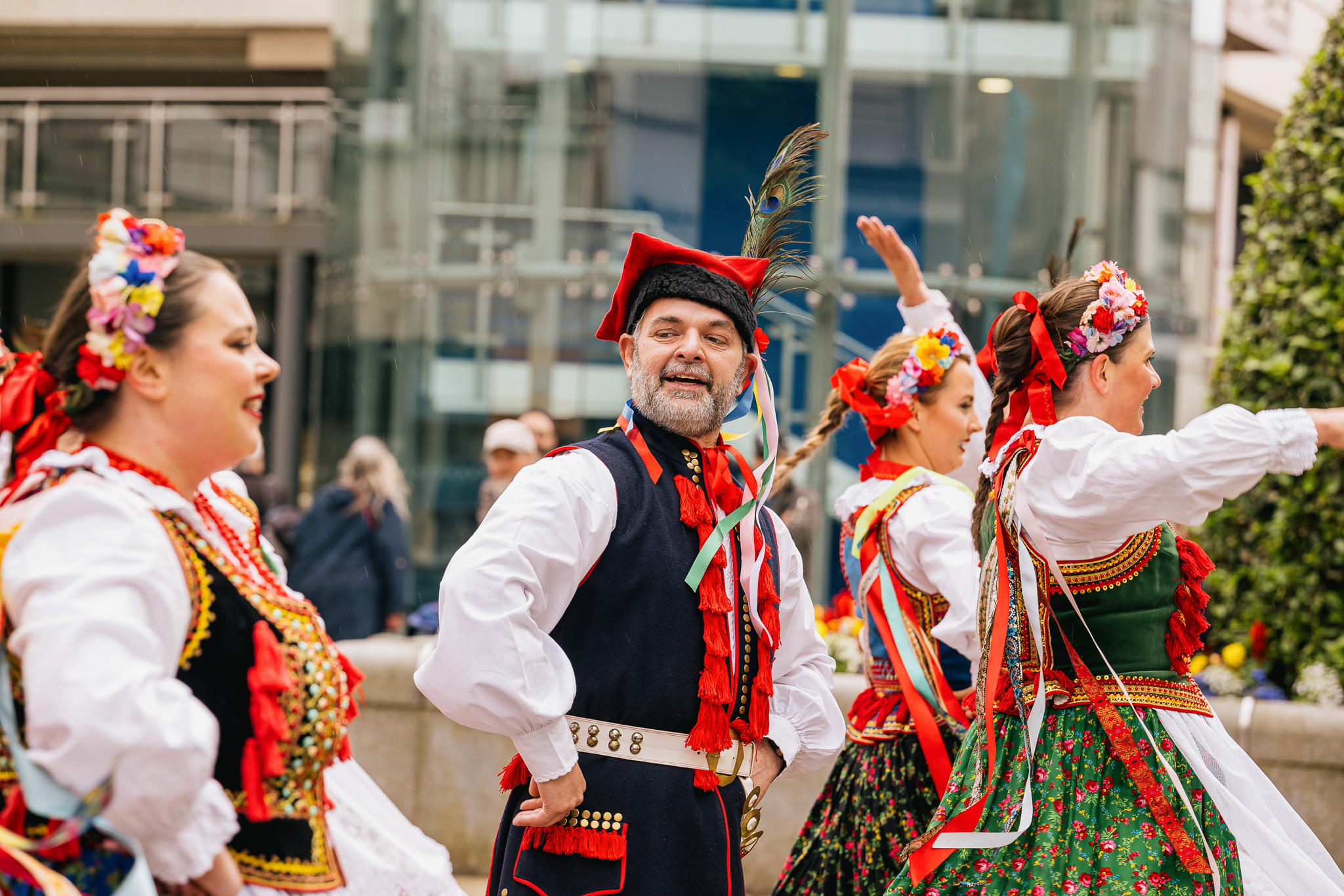 A group of people wearing colourful traditional folk costumes, with floral headpieces and ribbons, dance together outdoors in an urban setting. One man smiles and poses with his hand on his hip.