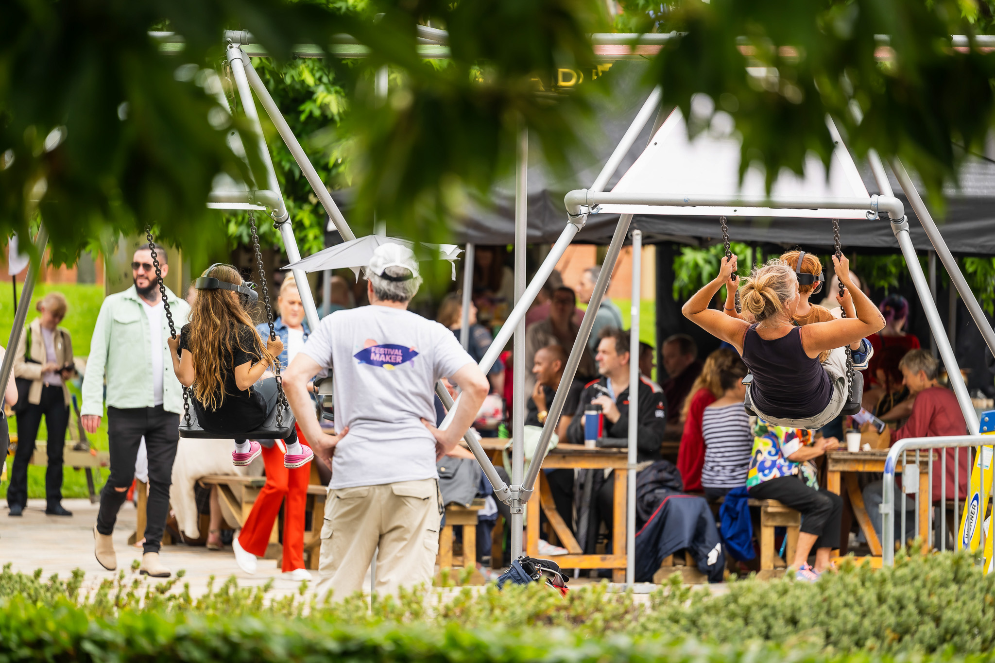 Two people swing on a large outdoor swing set while others sit and socialise at picnic tables under a canopy. The scene is lively, with greenery surrounding the area, and a man stands nearby watching.