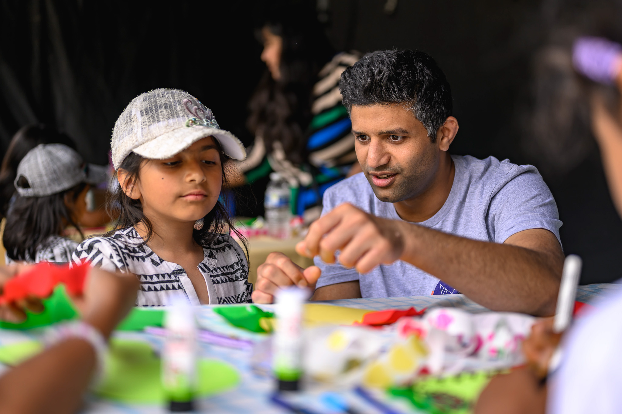 A man and a young girl sit at a table, engaged in an arts and crafts activity. The man is explaining something while the girl listens. Craft supplies and colourful paper are on the table, with other people blurred in the background.
