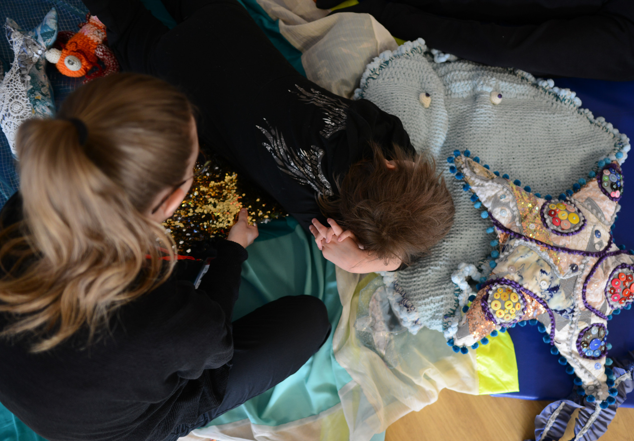 A child lies on colourful fabric with a starfish pillow, while a woman sits nearby, holding shiny sequins in her hand.