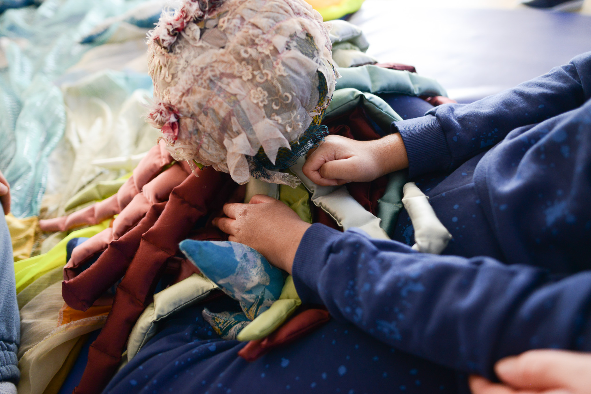 A child in blue clothing touches a colourful, textured fabric object adorned with lace, ribbons, and floral decorations, possibly part of a sensory or textile art activity.