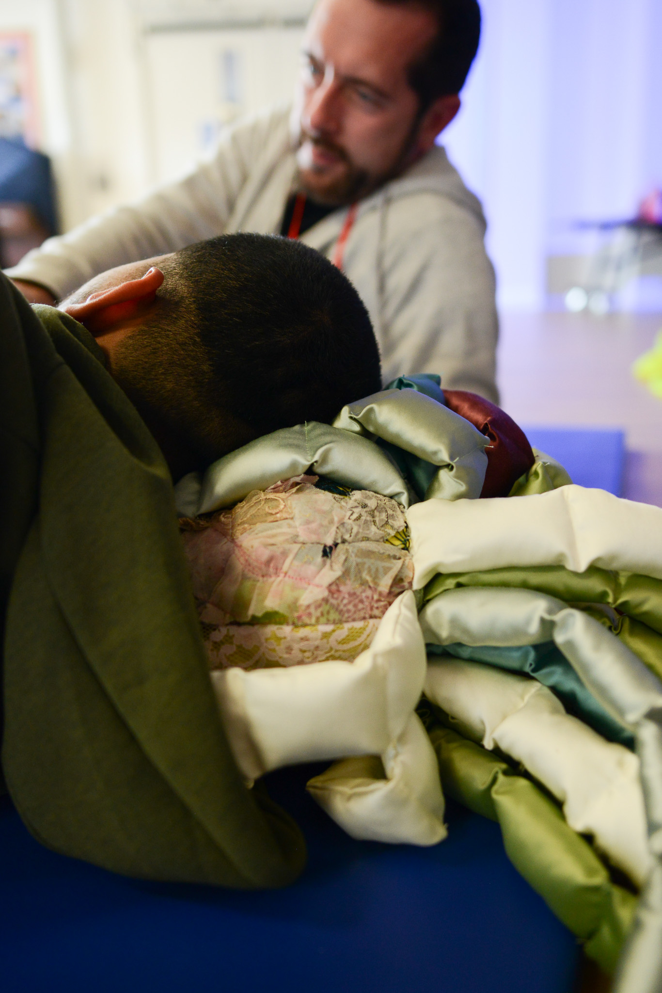 A man in a hoodie comforts a child wrapped in a colourful quilt, creating a warm, supportive atmosphere in a softly lit room.