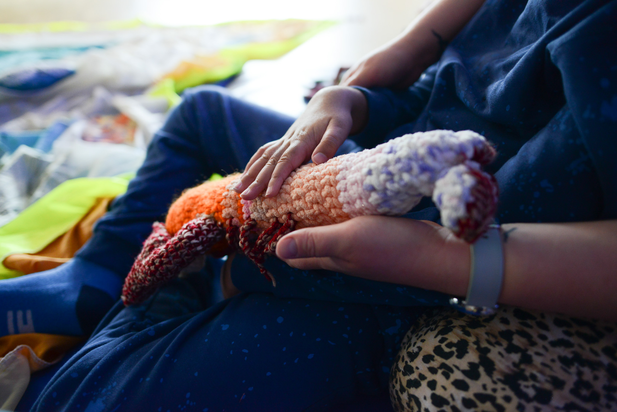 A person in blue clothing sits cross-legged, gently holding a crocheted soft toy shaped like a fox or animal, with their hand resting on it. Bright blankets are visible in the background.