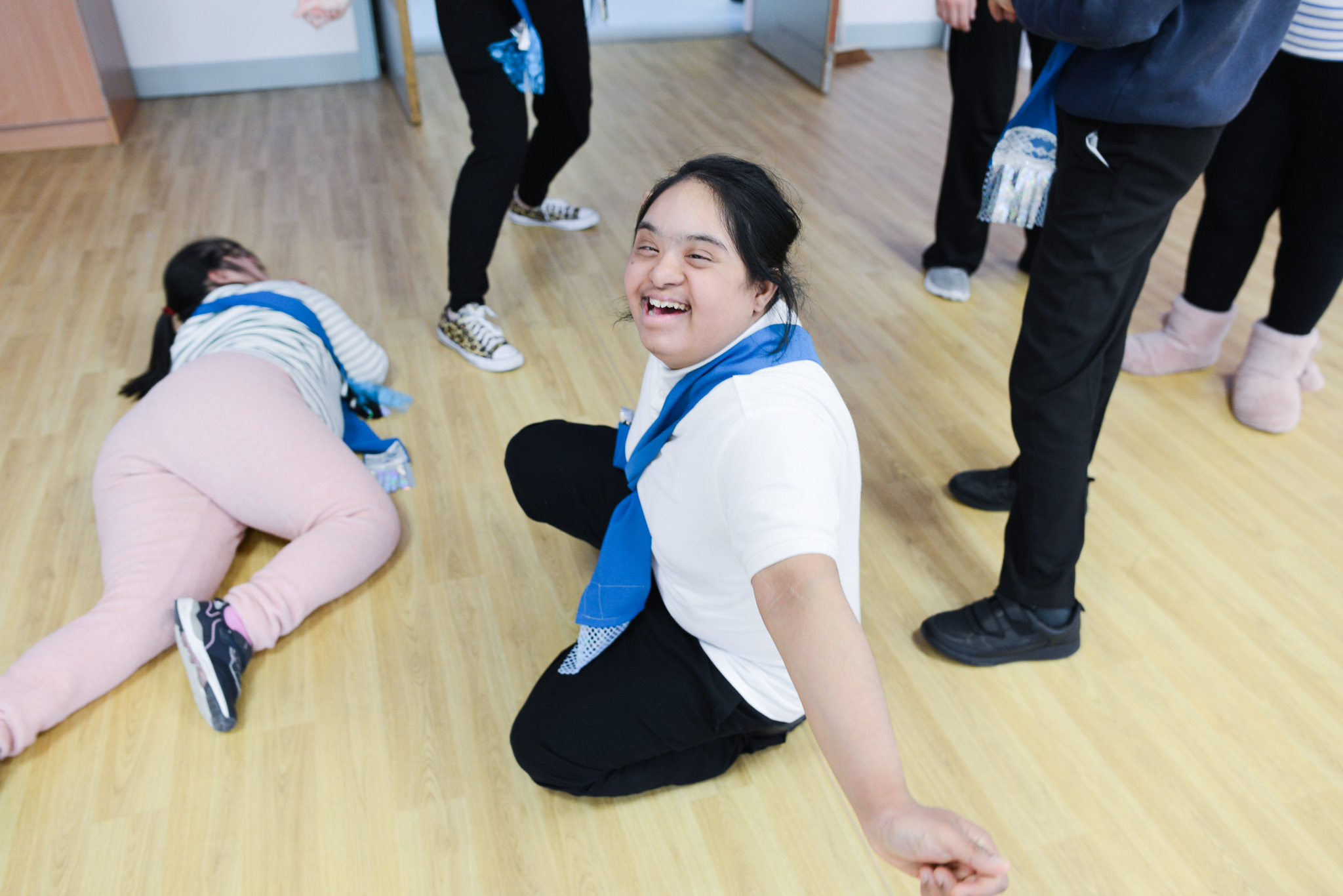 Smiling girl with a blue scarf sits on a wooden floor, surrounded by friends.