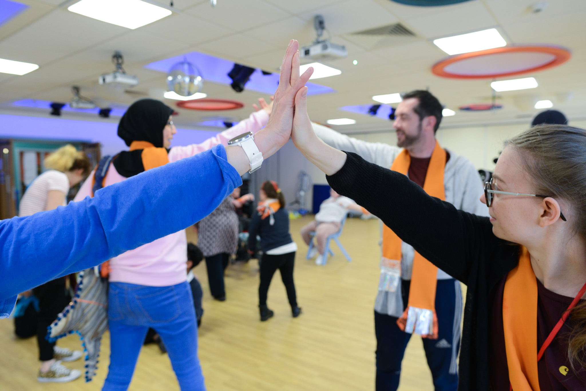 People in a bright room high-five, wearing orange scarves, engaging in a group activity.
