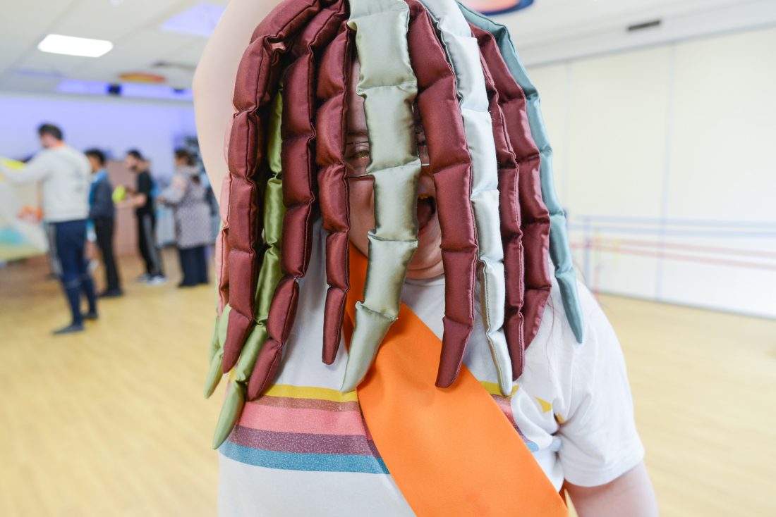 A person wearing a colourful costume with padded fabric strips covering their face stands indoors on a wooden floor. Other people are visible in the background, engaging in activities.
