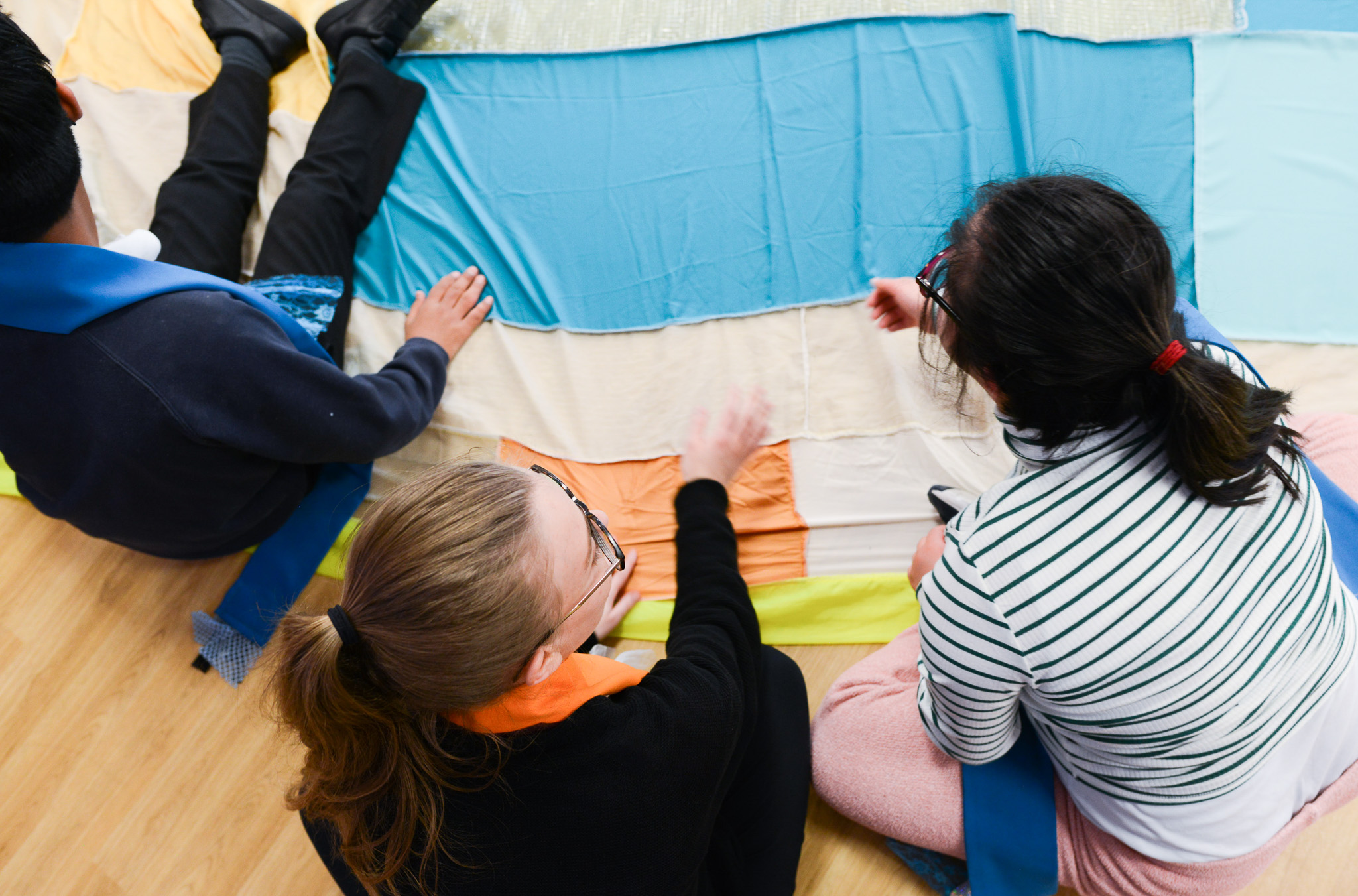 Three people sit on a wooden floor, arranging or touching colourful fabric pieces laid out in stripes. Two wear glasses and striped or solid shirts, and all appear engaged in a group creative activity.