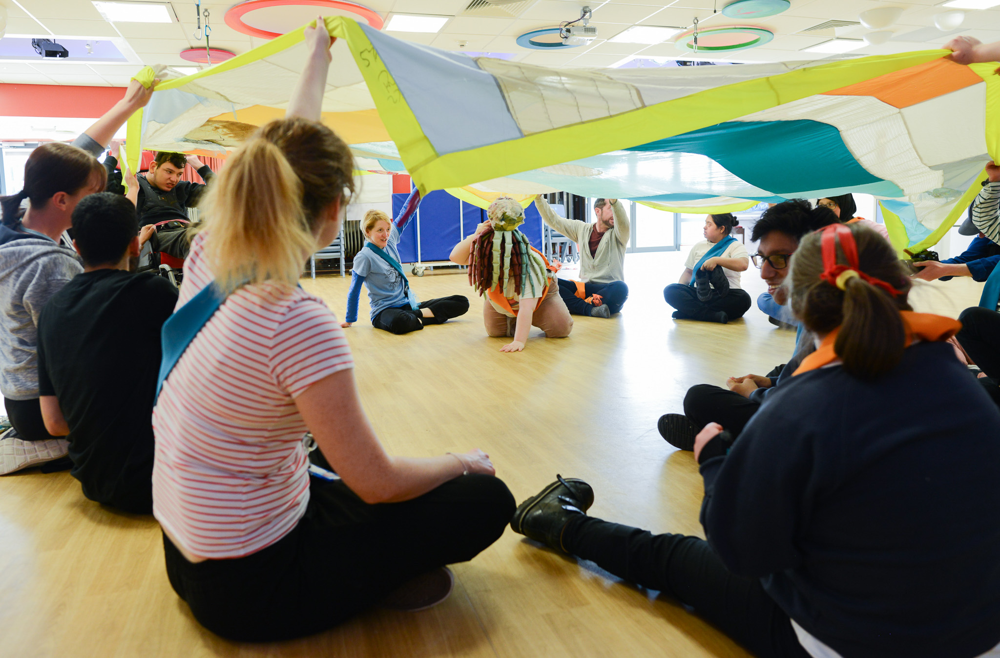 A group of people sit in a circle on a wooden floor, holding up a large colourful parachute above their heads, creating a playful and inclusive atmosphere indoors.
