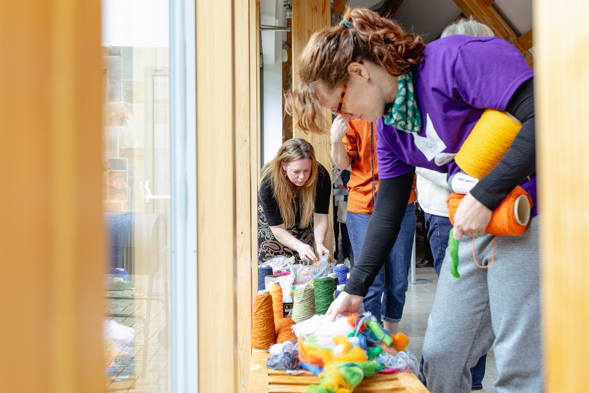 Two women sort through colourful yarn and wool on a wooden table indoors. One woman in a purple shirt is reaching for green wool, while another in the background kneels and looks at the materials. Natural light fills the space.