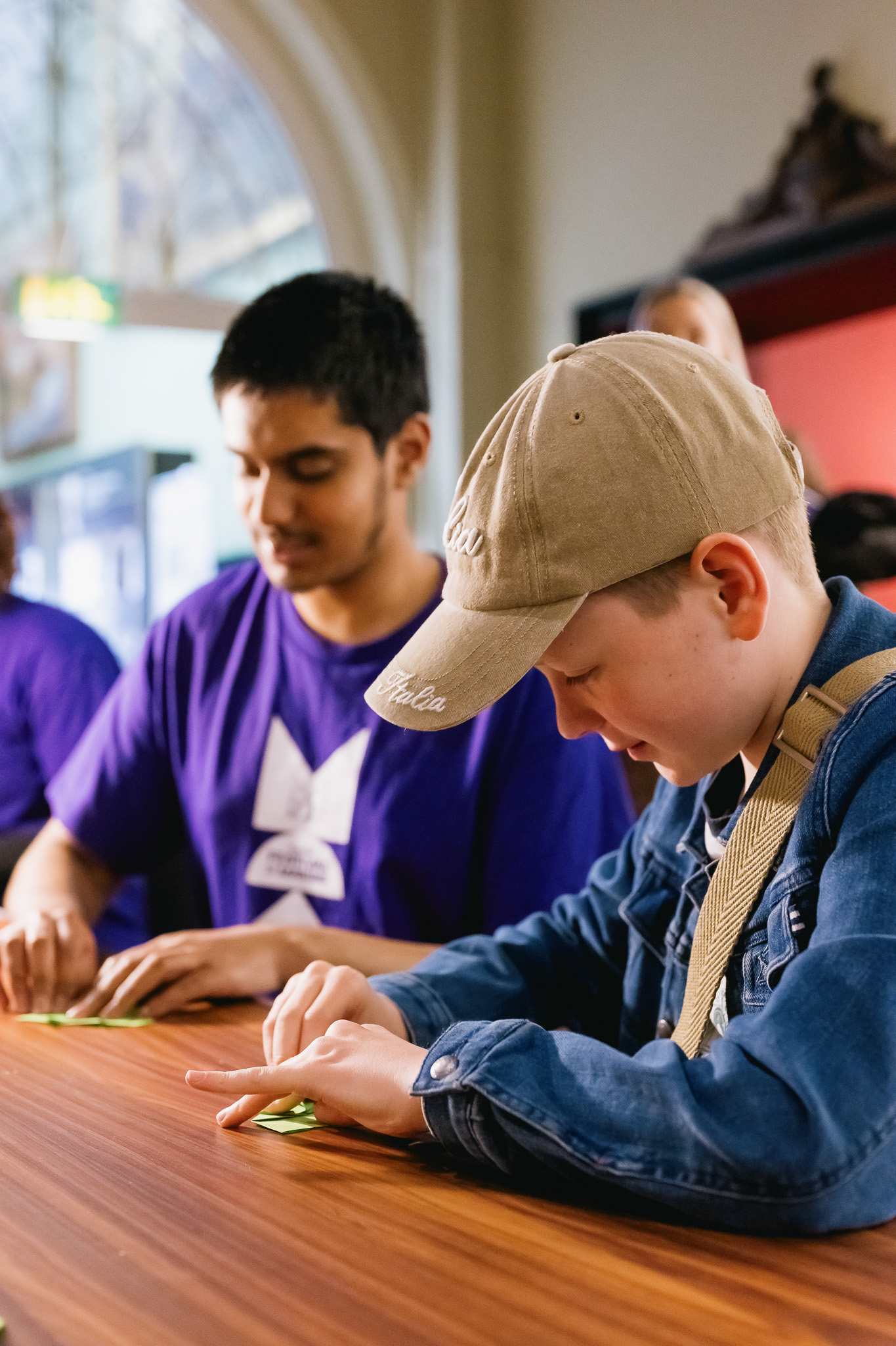 A boy wearing a beige cap and denim jacket folds paper at a table, while a person in a purple shirt assists beside him. They are participating in a practical activity indoors, under natural light.