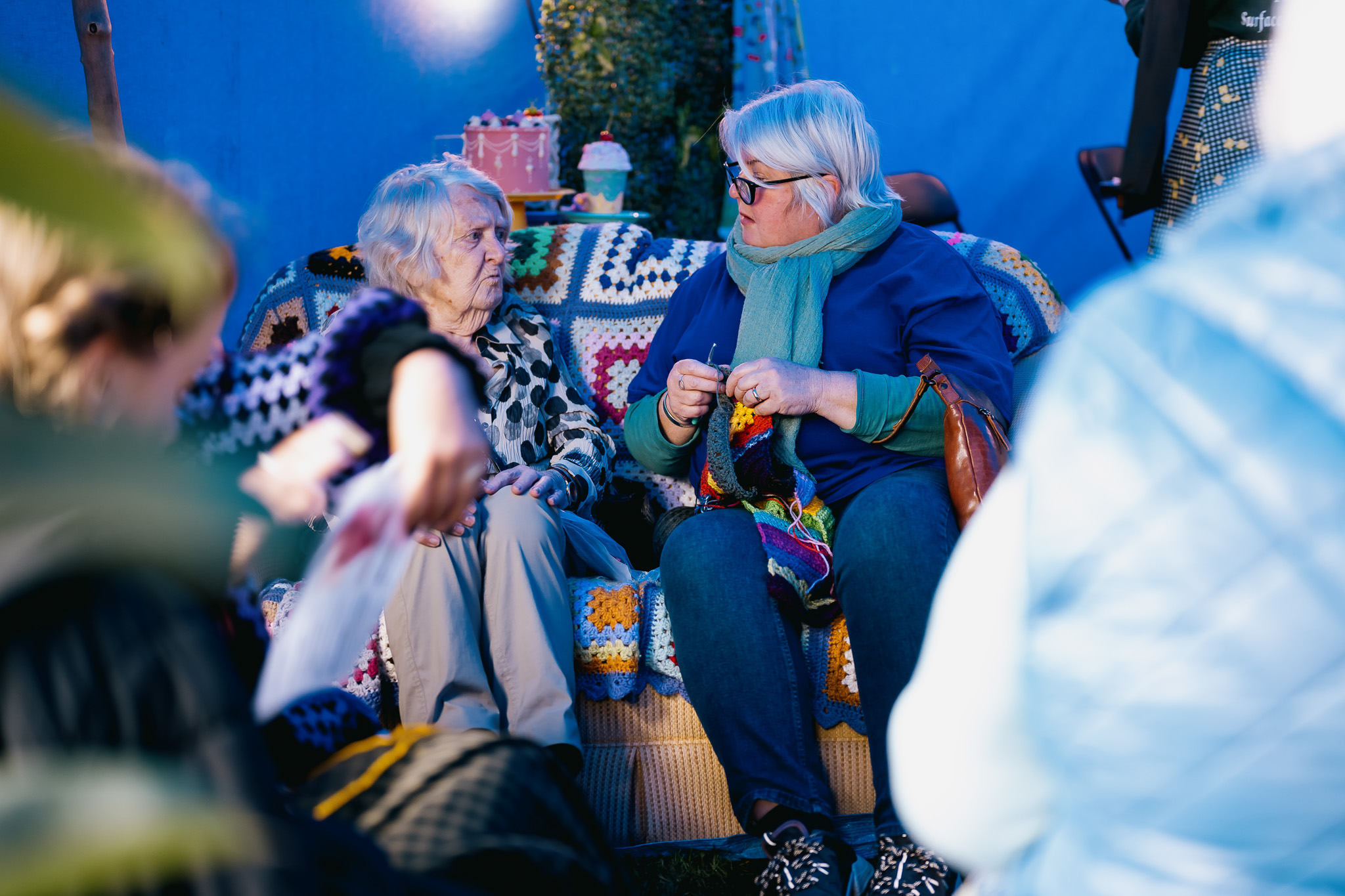 Two older women sit on a colourful crocheted blanket-covered sofa, engaged in conversation. One woman is knitting whilst the other listens. The scene is set outdoors with a blue backdrop and blurred figures in the foreground.