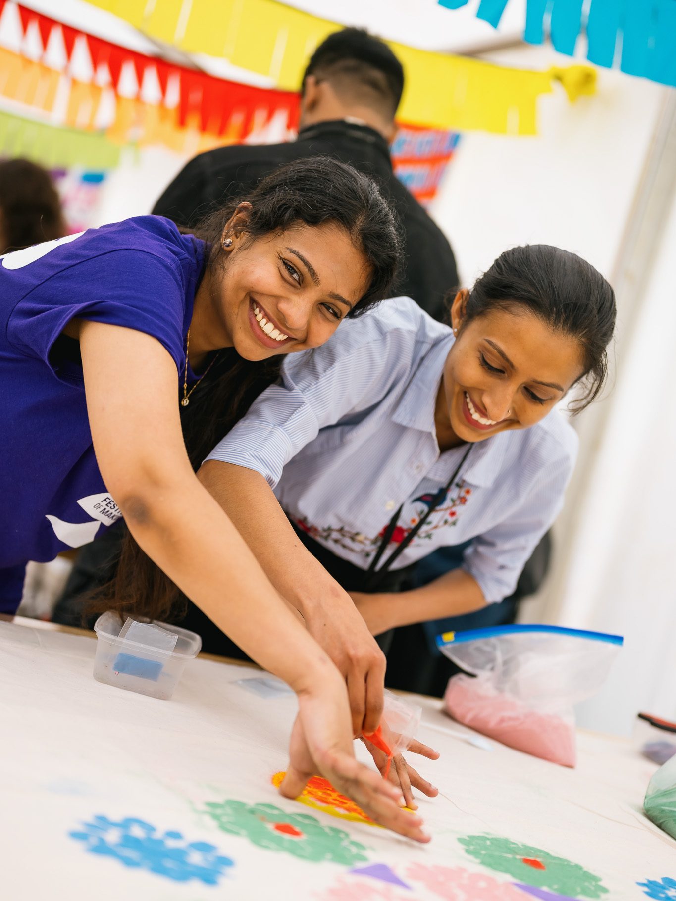 Two women smile as they create colourful art on a table covered with flower patterns, using coloured powders at a lively, festive indoor event decorated with vibrant hanging banners.