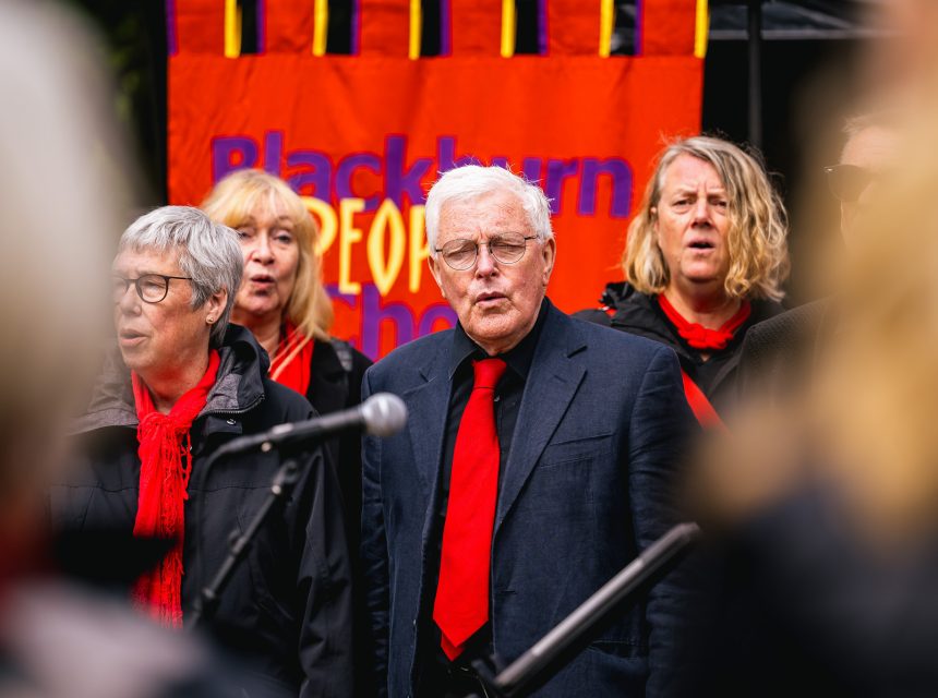 A choir group performs outdoors in front of a colourful banner. Members wear dark clothing with red scarves.
