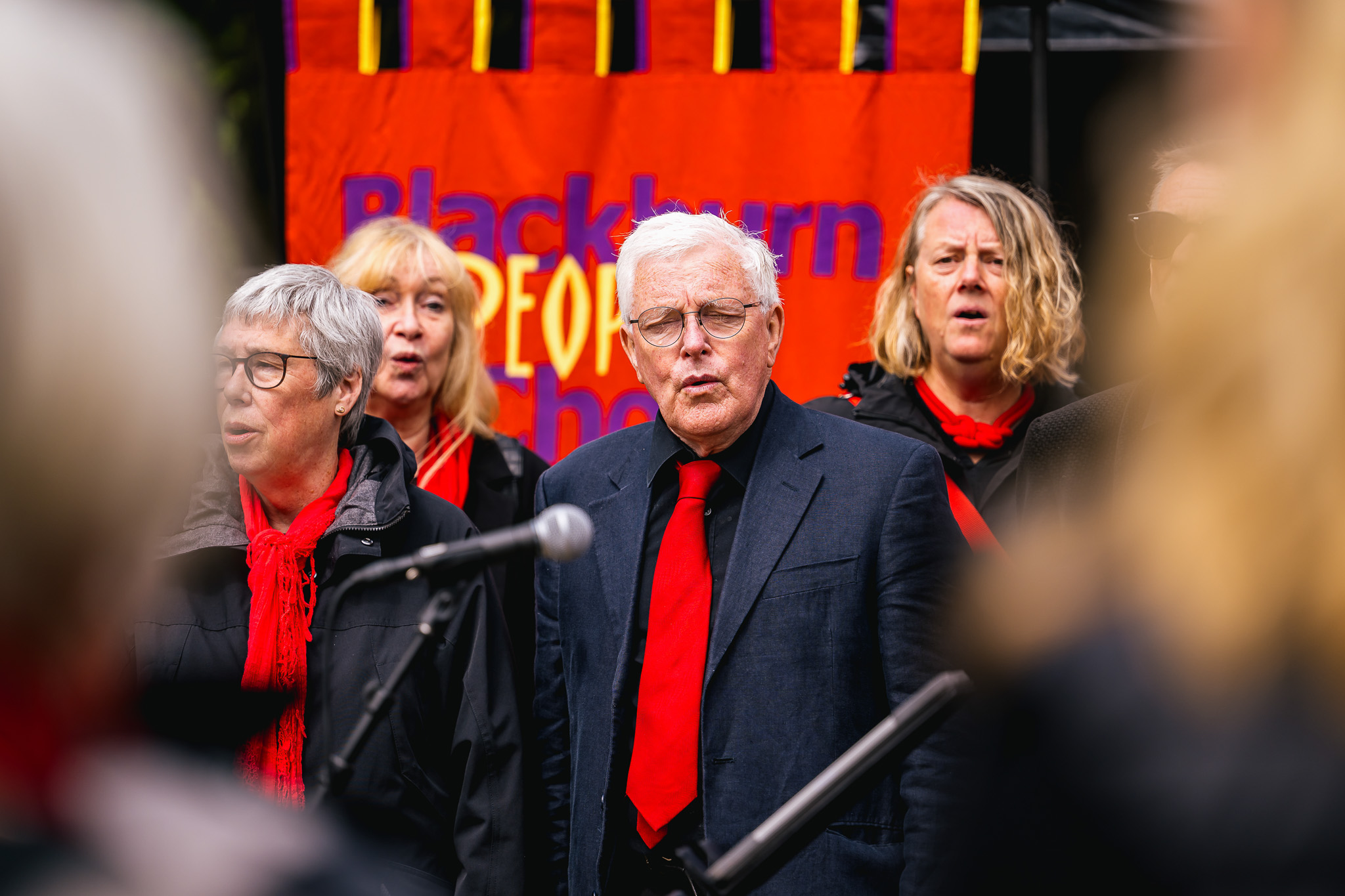 A choir group performs outdoors in front of a colourful banner. Members wear dark clothing with red scarves.