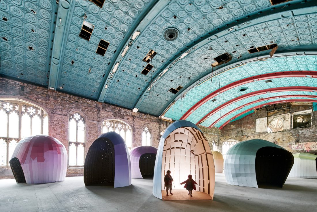 Two children walk out of a large, shell-like, cardboard structure inside a spacious hall with an ornate blue ceiling and arched windows. Several similar colourful pod installations are scattered around the room.