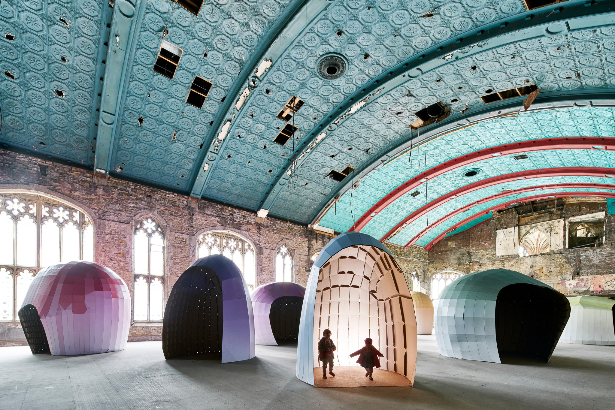 Two children walk out of a large, shell-like, cardboard structure inside a spacious hall with an ornate blue ceiling and arched windows. Several similar colourful pod installations are scattered around the room.