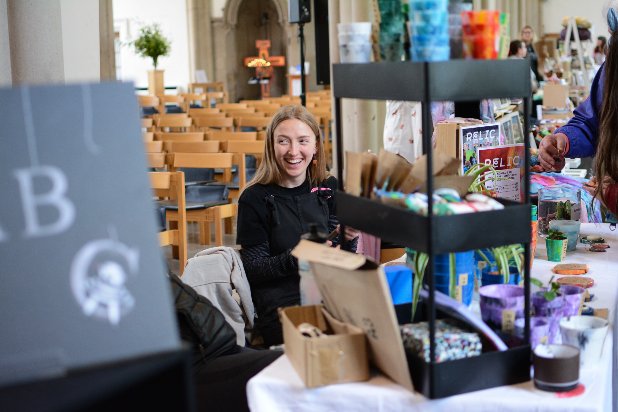 A woman with long blonde hair smiles whilst sitting behind a table displaying colourful crafts and products at an indoor market or fair, with rows of empty wooden chairs in the background.