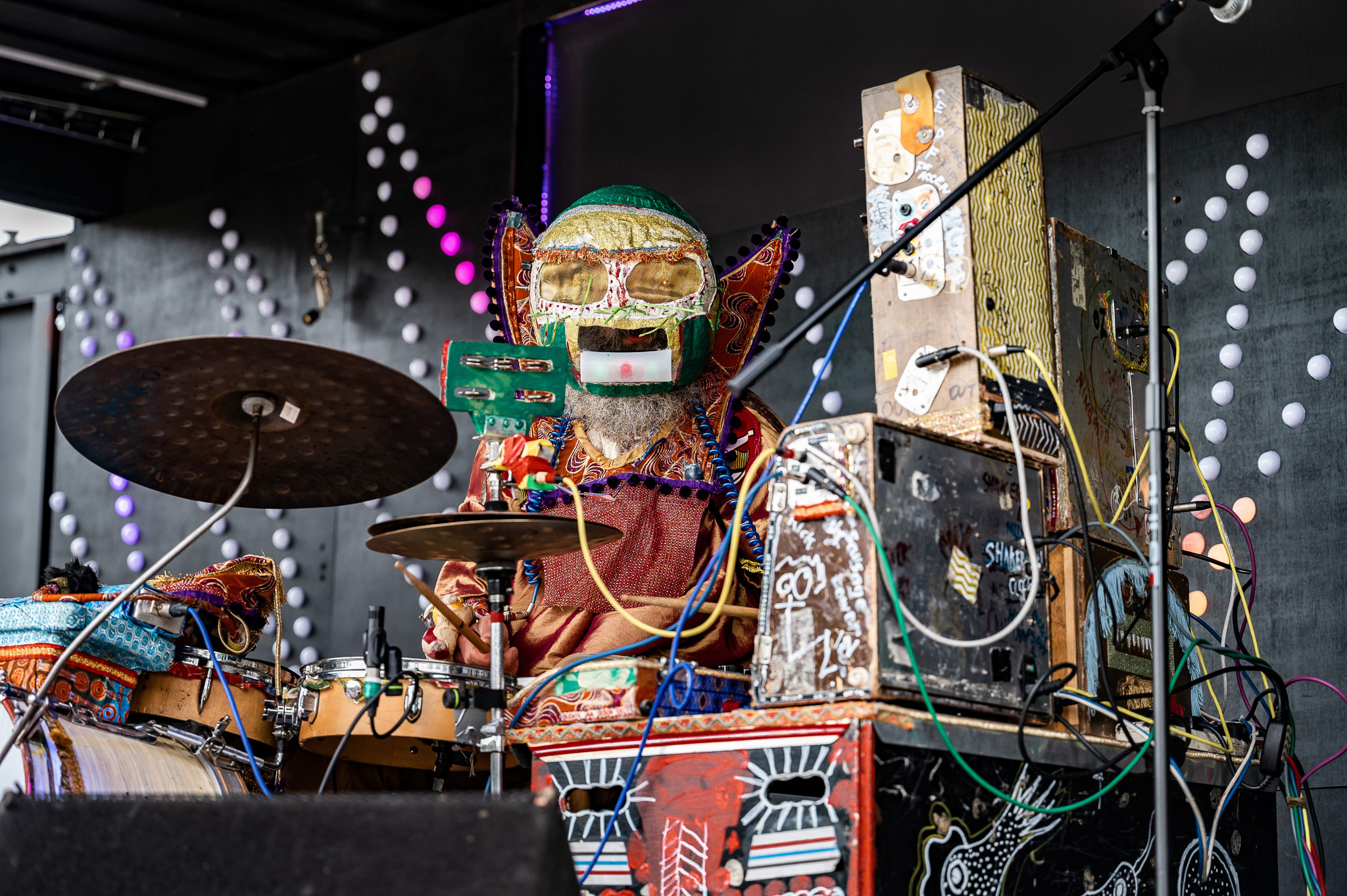 A person wearing a colourful, elaborate costume and a decorative mask plays a drum kit on stage, surrounded by musical equipment and vibrant, patterned decorations.