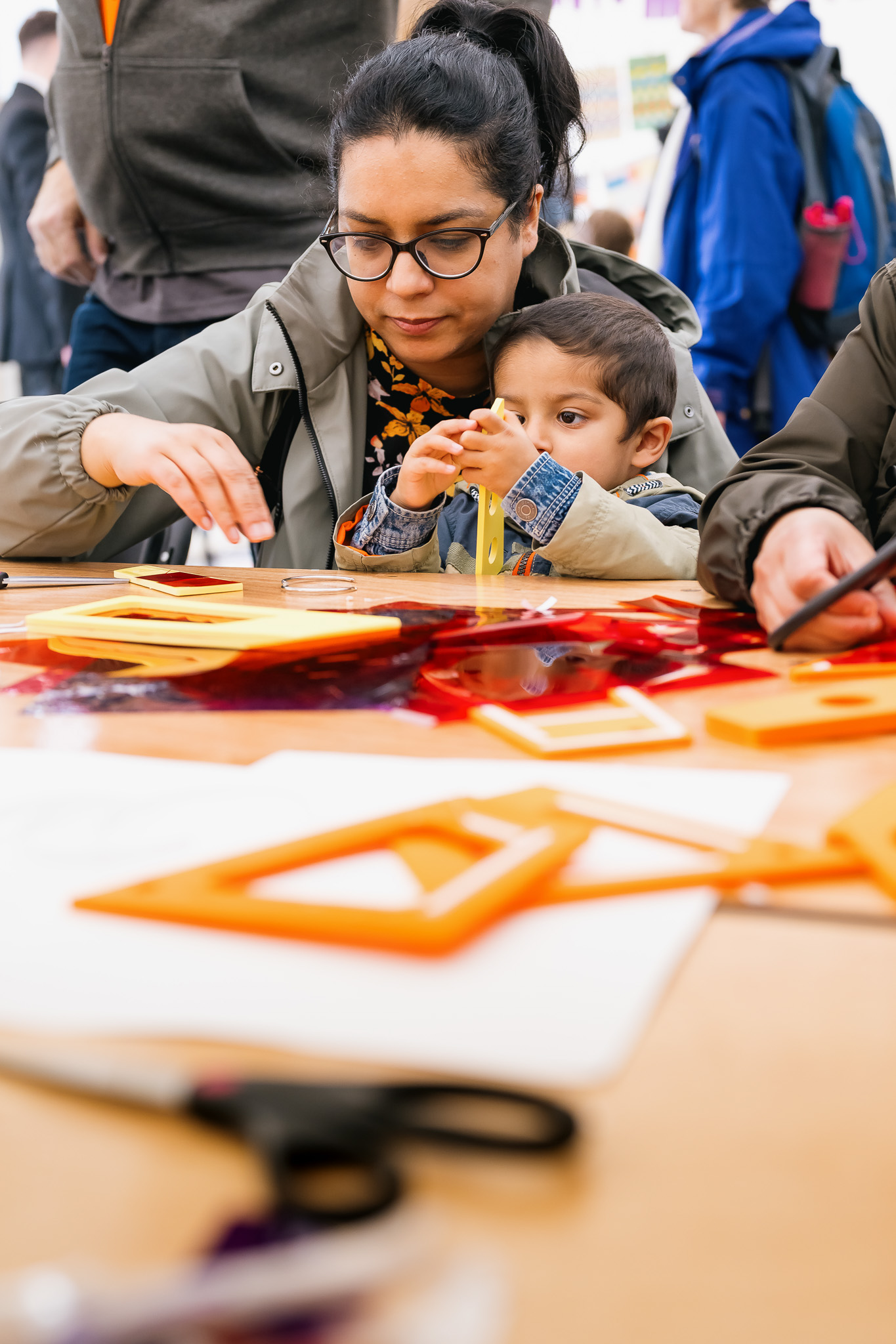 An adult and child engaged in a workshop, surrounded by colourful art supplies on a table.