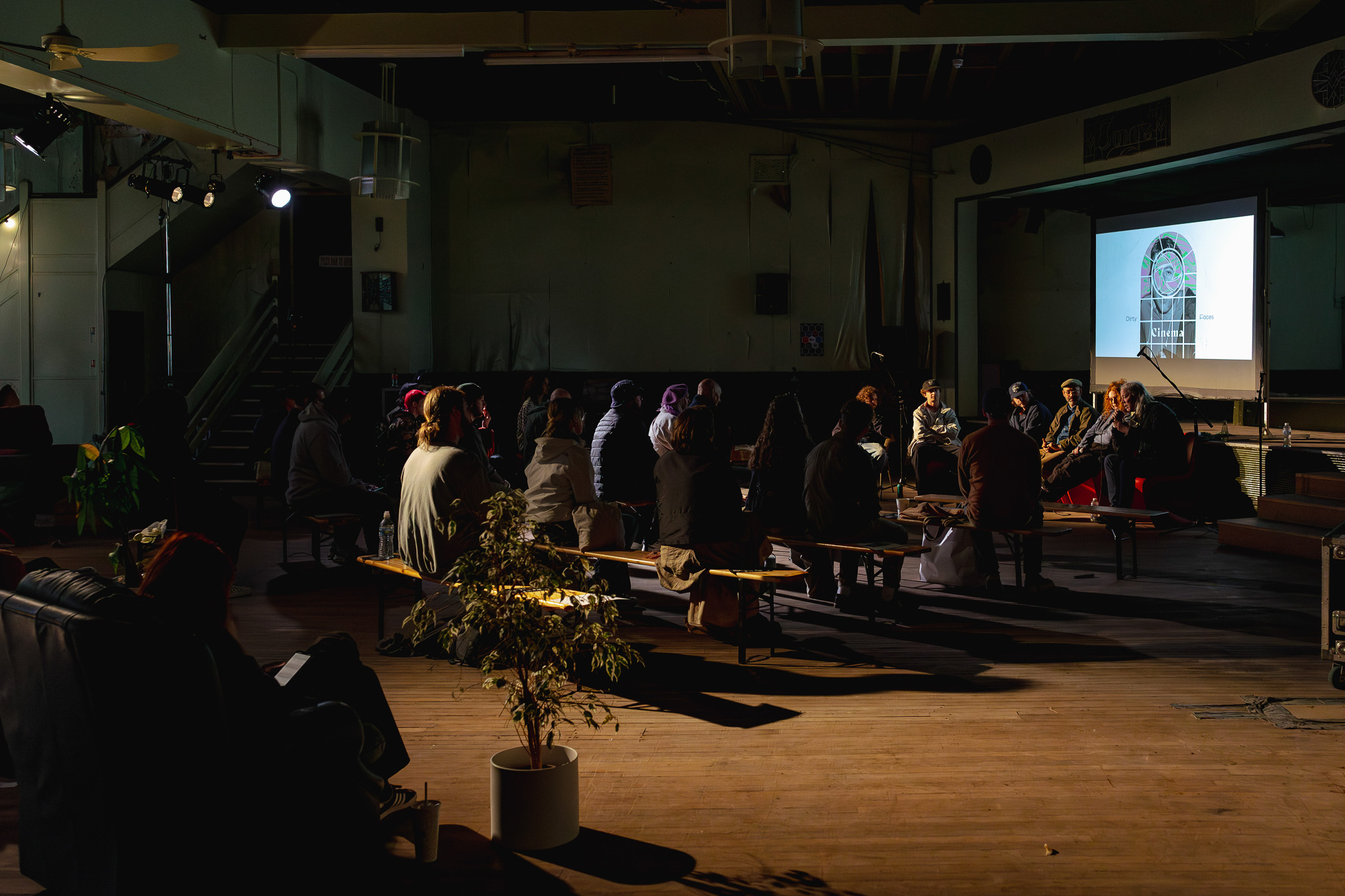 A group of people sits on sofas and chairs in a large hall, watching a panel talk. The room has dim lighting, wooden floors, and eclectic furniture.