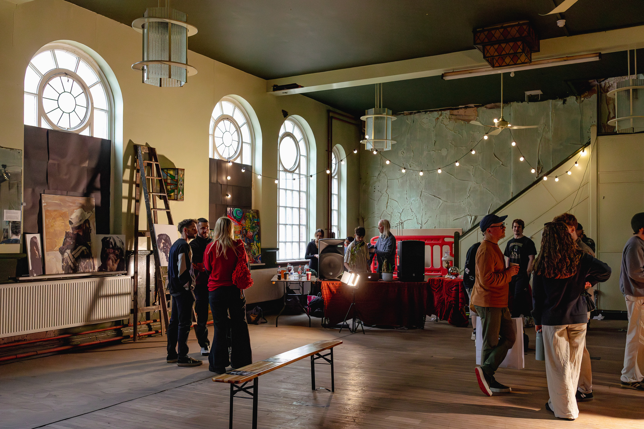 People gather and chat in a spacious, sunlit room with large arched windows, fairy lights, and various artworks displayed on the walls. A ladder and art supplies are visible, creating an artistic, informal atmosphere.