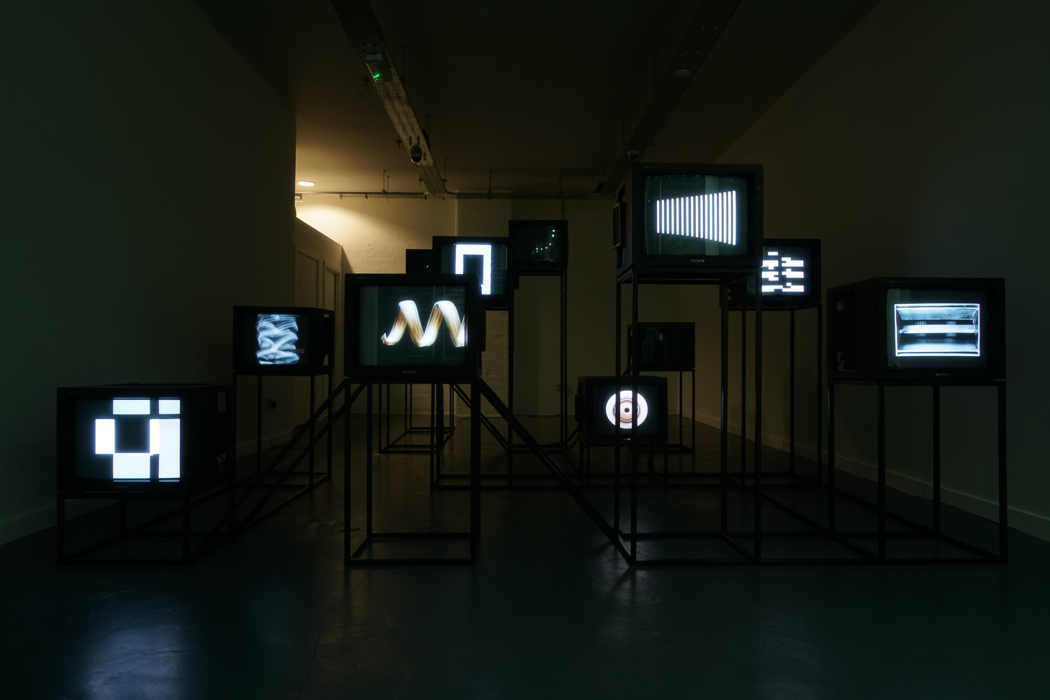 A dark room with multiple TV screens on metal stands, each displaying abstract black-and-white patterns and shapes. The screens are arranged at different heights and angles, creating a modern art installation.