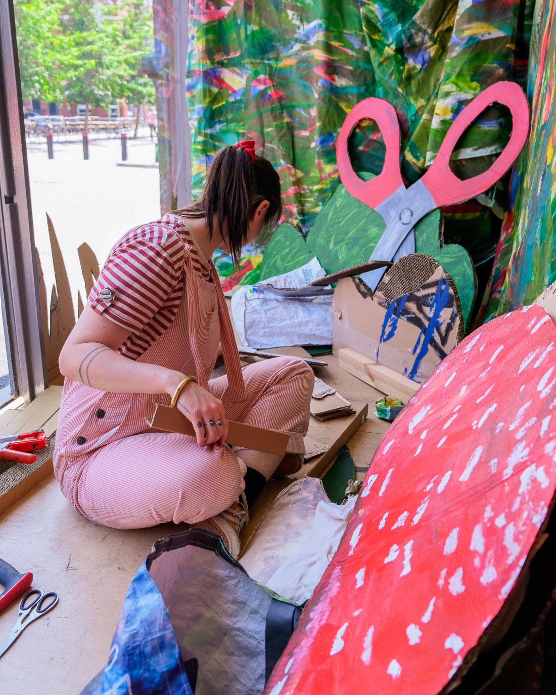 A person in pink dungarees sits on the floor next to a window, assembling colourful, large cardboard props, including a red mushroom and giant scissors, surrounded by painted cardboard scenery.