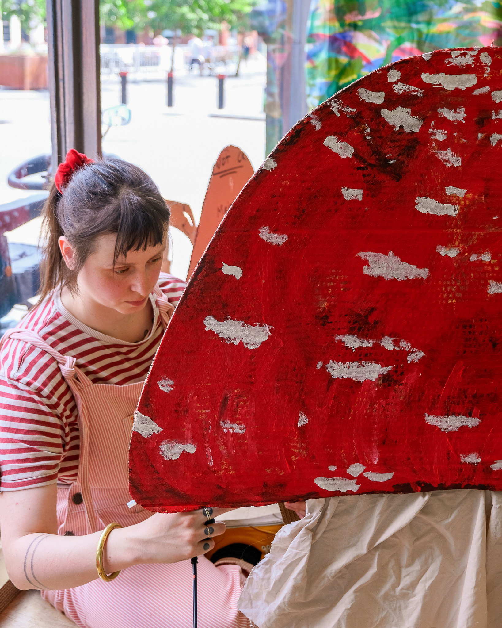 A person in pink dungarees and a red-and-white striped shirt paints a large red toadstool prop with white spots indoors, near a window with trees and parked cars visible outside.