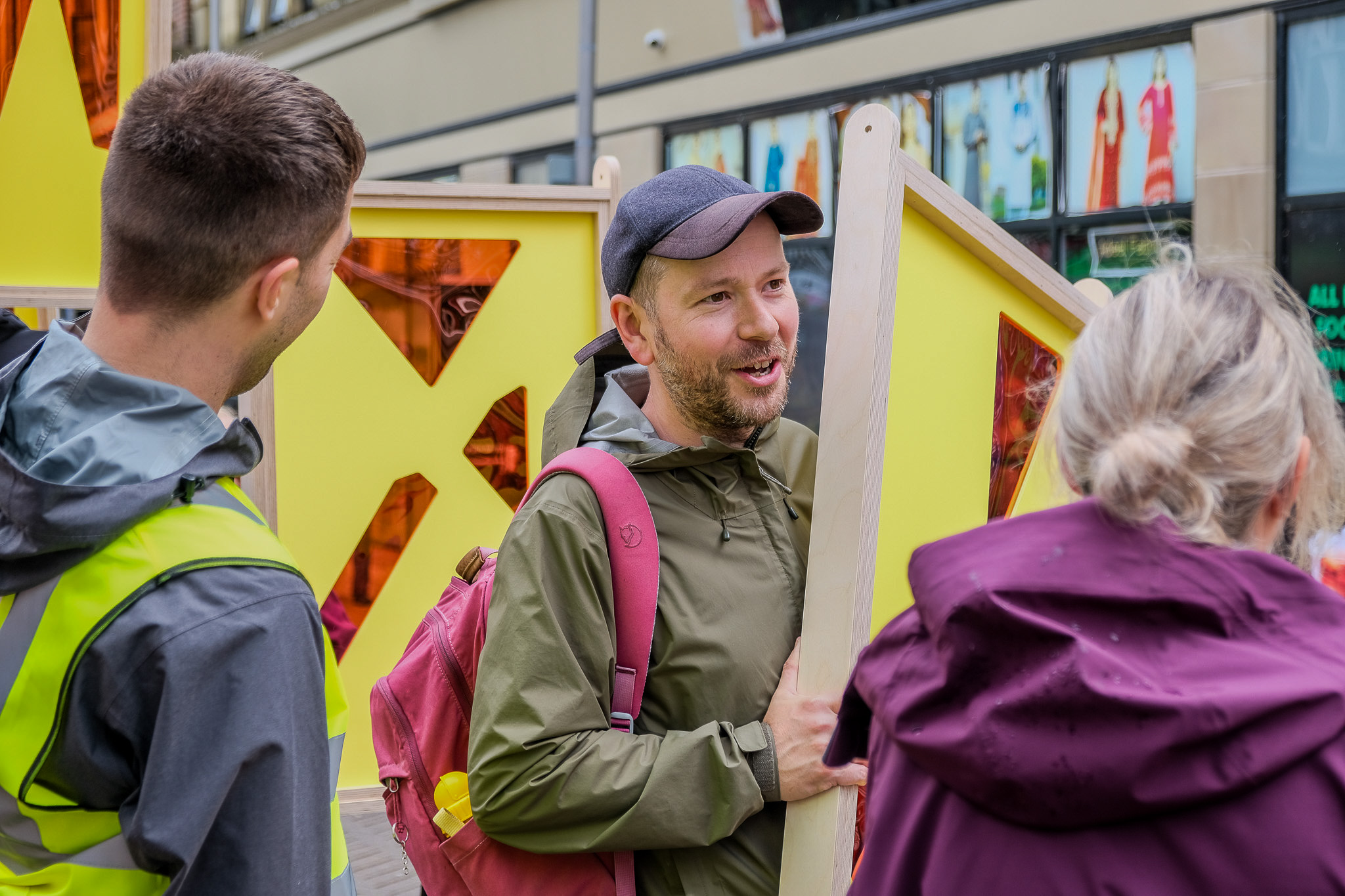 A group of people stand outside, engaged in a lively conversation. One person holds a bright yellow frame.