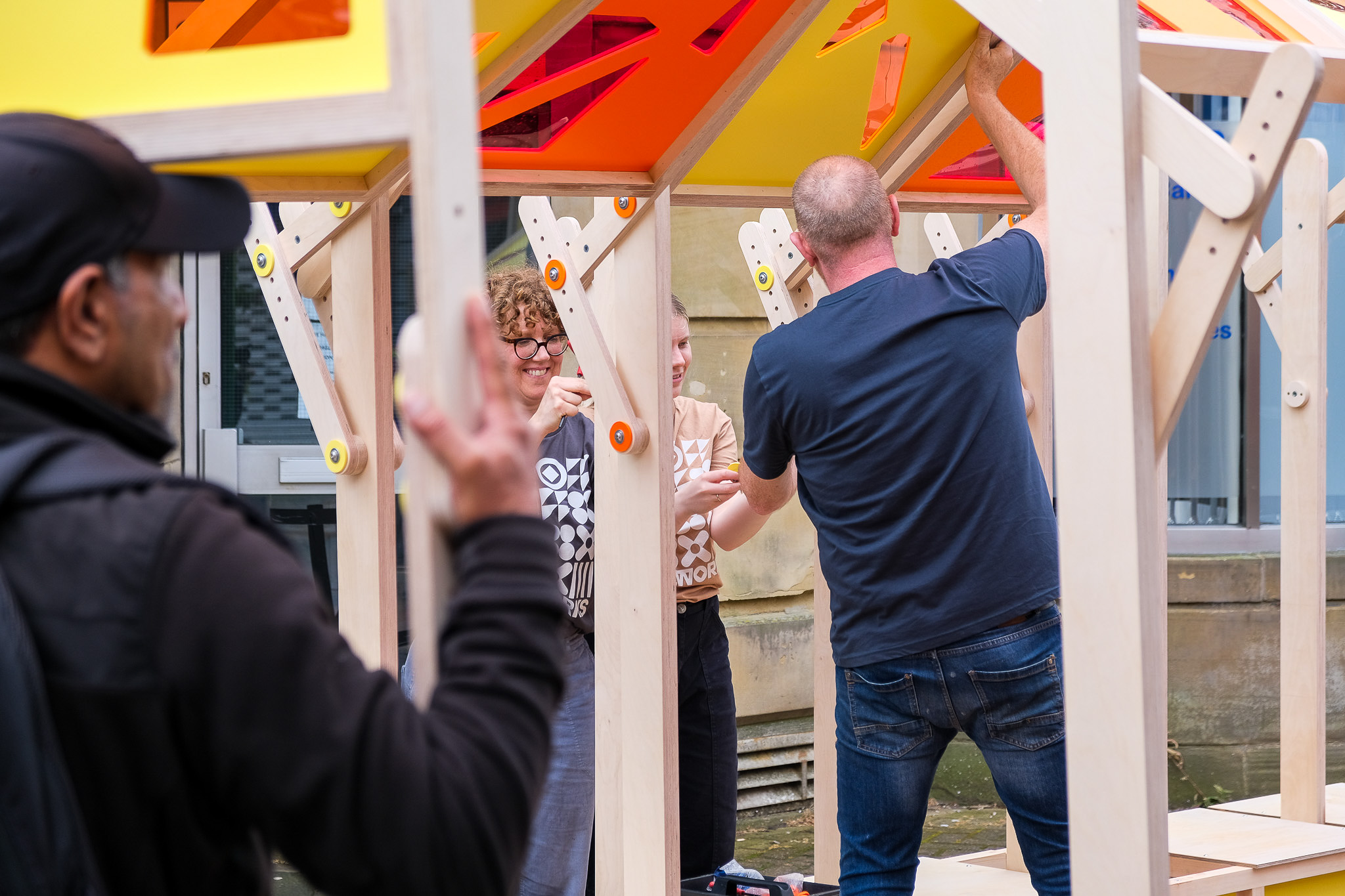 Several people work together assembling a wooden structure with bright yellow and orange panels outdoors. One person faces away, adjusting parts, whilst others hold pieces and converse, focused on the project.