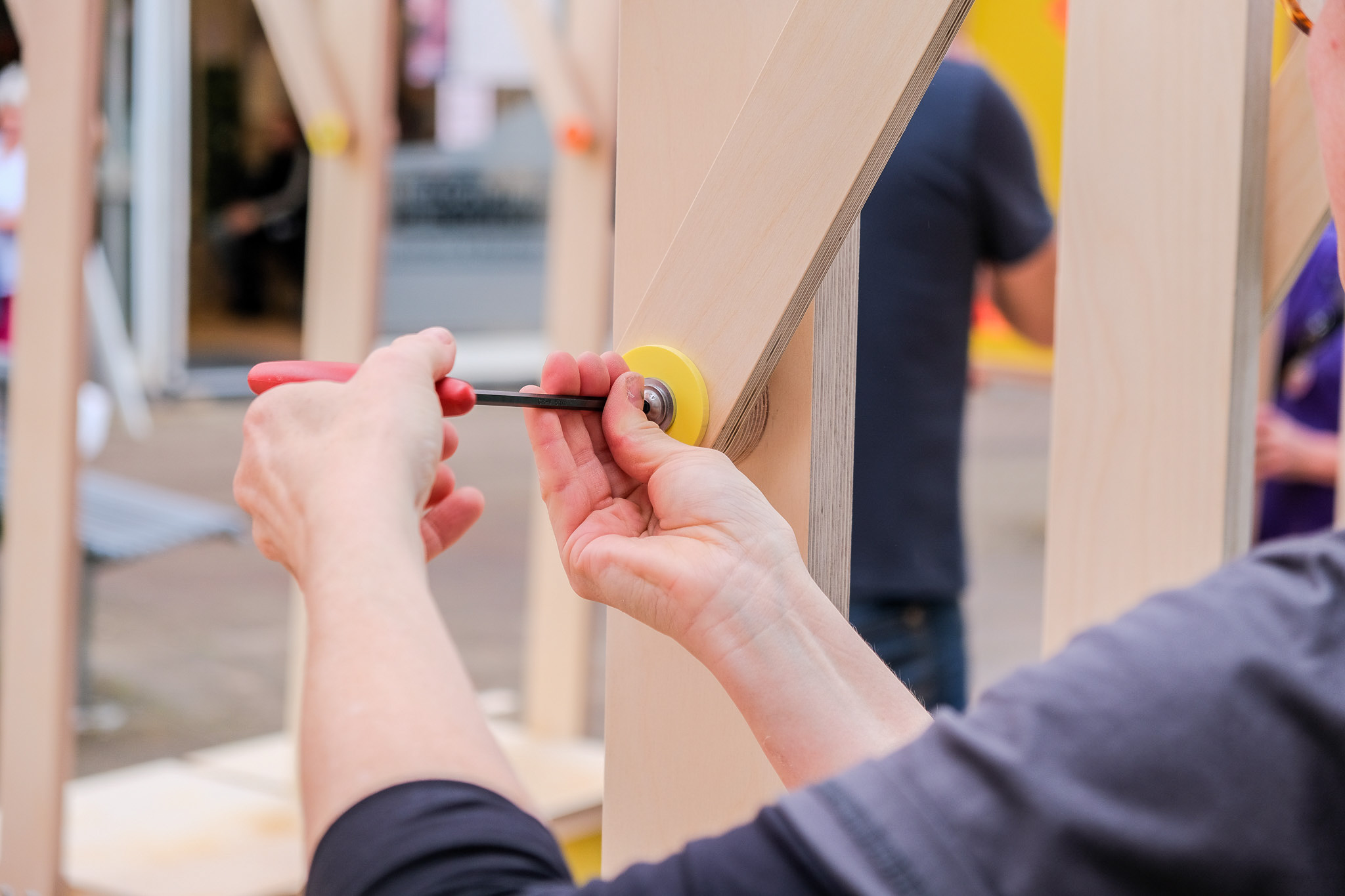 Hands tighten a bolt with a screwdriver on a wooden structure, outdoors.
