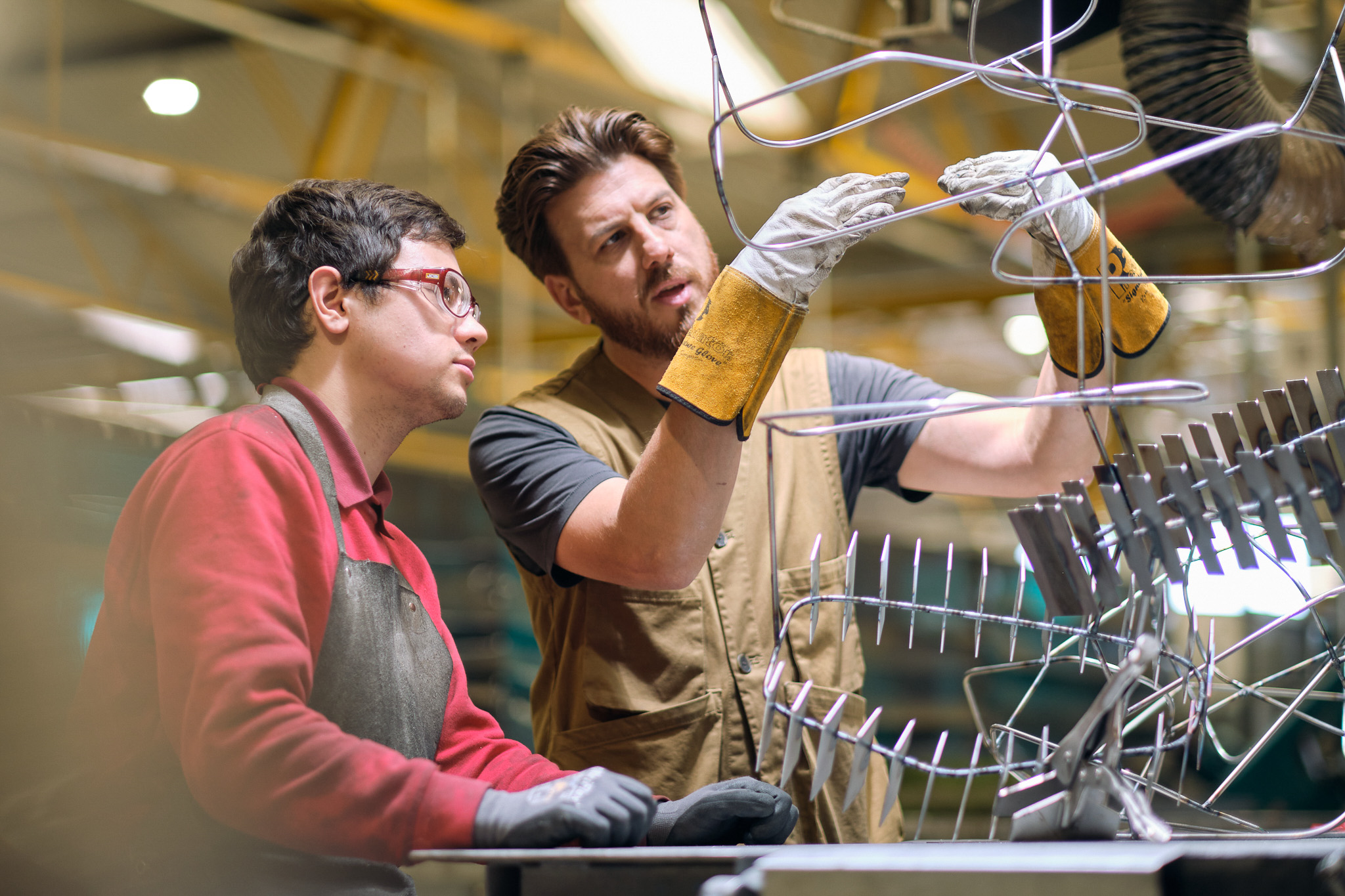 Two factory workers wearing protective gloves and aprons work together on assembling or inspecting a metal structure in an industrial setting. One worker gestures and explains whilst the other observes attentively.
